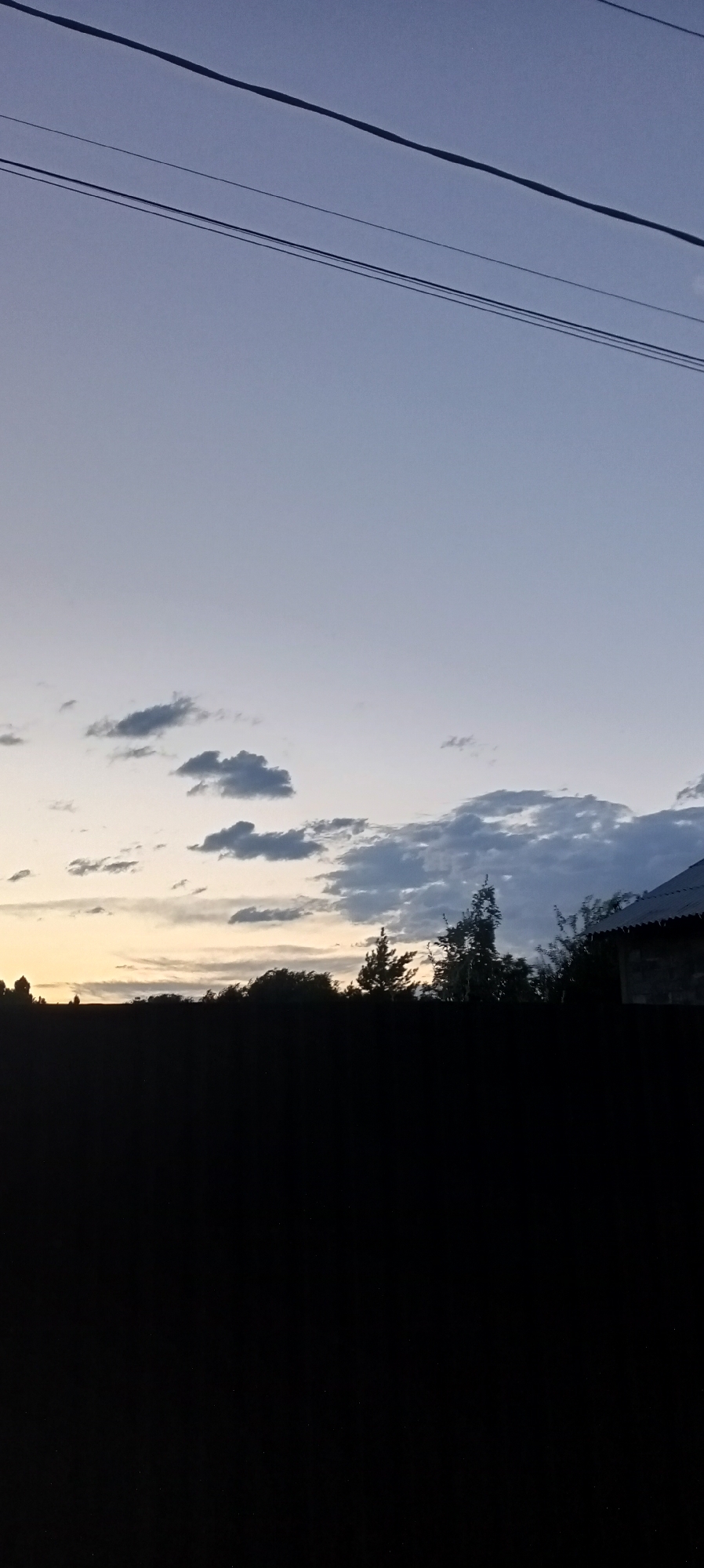 The image depicts a twilight sky with a gradient of pale blue and faint yellow hues, suggesting either dawn or dusk. Several thin, dark clouds are scattered across the sky, some appearing wispy and others more defined. Three parallel, thin, dark lines, likely power lines, traverse the upper portion of the frame.

Below the sky, a dark silhouetted landscape is visible. This landscape appears to be a treeline with varying heights of foliage, including what looks like coniferous and deciduous trees. To the right, a portion of a building with a sloped, dark roof is visible, partially obscured by the trees. The bottom half of the image is entirely black, indicating an area of deep shadow, possibly foreground or a dark fence.

The scene is serene and appears to be an outdoor setting. No people are visible in the image, and there are no obvious actions or interactions occurring. The location is stated to be in the city of Romanovka, Kyrgyzstan.
