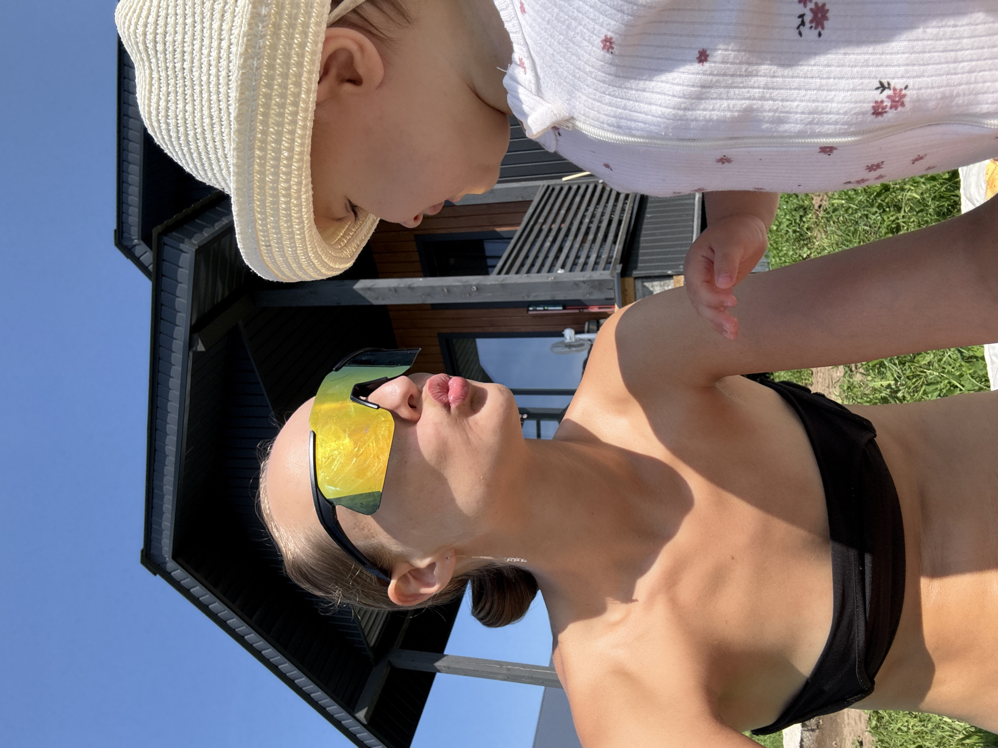 A woman with blonde hair pulled back, wearing a black bikini top and large yellow-tinted sunglasses, makes a kissing face towards a baby. The baby, wearing a white sun hat and a white onesie with a floral pattern, looks at the woman. They are outdoors with a modern wooden house with dark siding and a dark roof in the background. The sky is clear and blue. Green grass is visible in the foreground. The location is in the village of Podshivalovo, Russia.