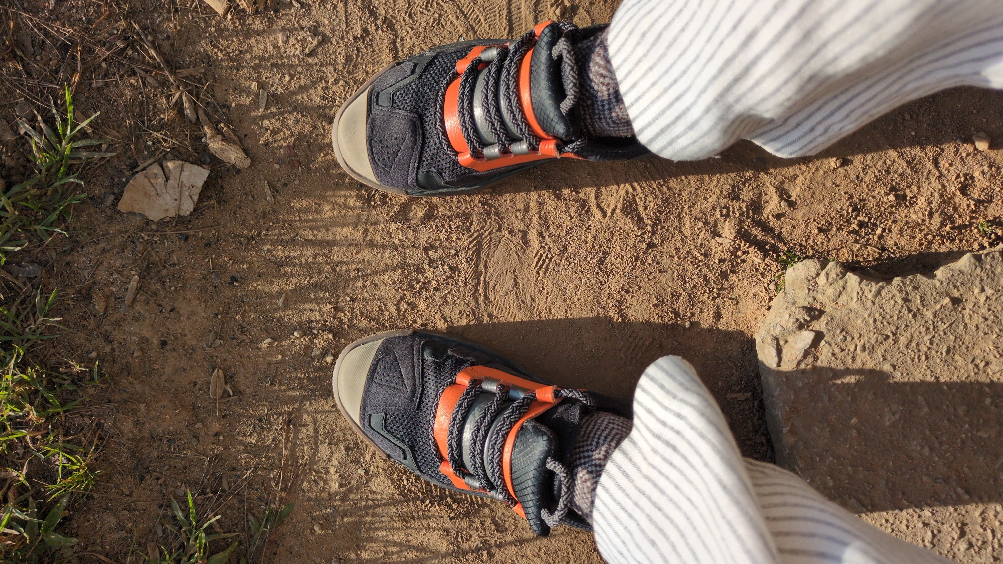 This is a top-down, eye-level shot of a person wearing black hiking shoes with orange accents and gray laces, standing on a dirt path in Da Lat, Vietnam. The path is composed of light brown dirt, with scattered small stones and dry leaves. Green grass and foliage are visible in the upper portion of the frame, suggesting an outdoor, possibly natural setting within the city. The person's lower legs are visible, covered by striped white pants. Shadows from the shoes and surrounding vegetation are cast across the path.