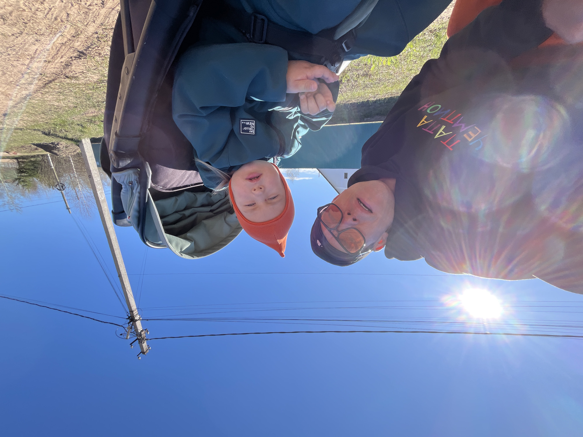 Captured outdoors in daylight, the image portrays a selfie featuring two individuals against a bright blue sky with visible power lines and the sun's glare. A young child, wearing an orange beanie and a teal jacket with a patch that reads "SAY HELLO! TO NEW***," is positioned within what appears to be a baby stroller or carrier. Next to the child, an adult is visible, wearing sunglasses and a black hoodie, part of which bears a text design. An electric pole and trees are in the background. The location is Chaikovsky, Russia.