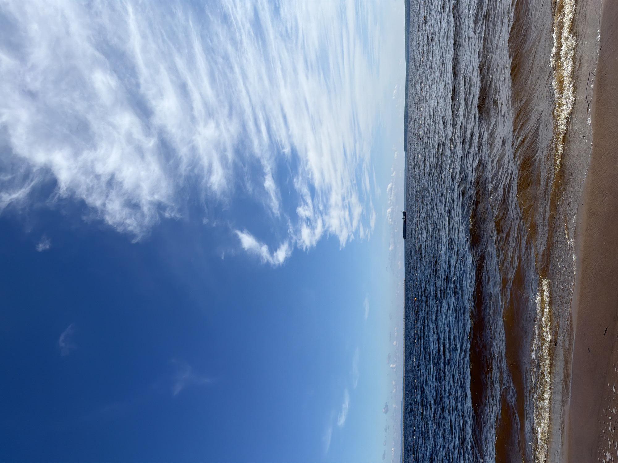 The image, when viewed in its intended landscape orientation, presents a wide vista of a large body of water and the sky above it, with a narrow strip of sandy shore along the right edge.
The sky occupies the upper portion of the frame, showcasing a beautiful gradient from a rich, deep blue in the upper left to a lighter, paler blue closer to the horizon. Wispy, streaky white clouds, likely cirrus, stretch across the top and upper-middle sections, particularly prominent on the left side, adding texture and a sense of movement to the expansive blue.
Below the sky lies a vast expanse of water, which appears dark blue-grey, suggesting depth. Its surface is covered with visible ripples and small waves, reflecting hints of sunlight in places. Towards the right side of the frame, these waves gently meet a light brown, sandy shore, where a line of white foam marks the immediate waterline. A small piece of natural debris, possibly a twig, is visible on the sand.
Along the distant horizon, a faint, dark line indicates a faraway landmass or coastline. A single, small, dark boat or ship is discernible on this distant horizon, slightly to the left of the center. Further into the middle ground of the water, a series of small, light-colored buoys or markers can be seen, suggesting a designated area or navigational path.
The scene depicts a calm and sunny day, likely mid-morning or afternoon, with fair weather indicated by the clear blue sky and high-altitude clouds. The overall impression is one of serene natural beauty and vast open spaces. Given the location context of Chaikovsky, Russia, this is likely a view of the Kama River or the Votkinsk Reservoir. There is no visible text in the image.