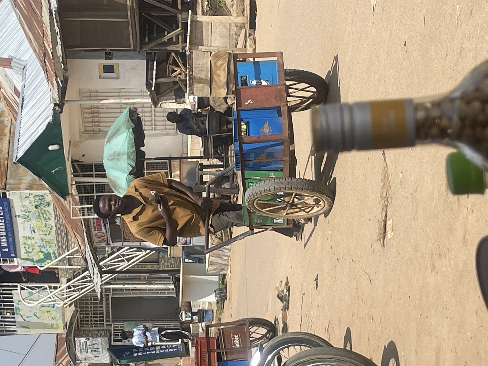 In an outdoor market scene in Abuja, Nigeria, a man is pushing a cart loaded with blue and red crates. The cart has two large spoked wheels. The man, wearing a brown short-sleeved shirt and dark pants, is looking at his mobile phone while holding the cart's handles. In the background, several market stalls with corrugated metal roofs are visible. One stall has a green umbrella set up. Other people are present in the background, including a person standing near a staircase and another seated behind a stall. The ground is dusty and uneven. The scene captures a typical street market environment in Abuja.
