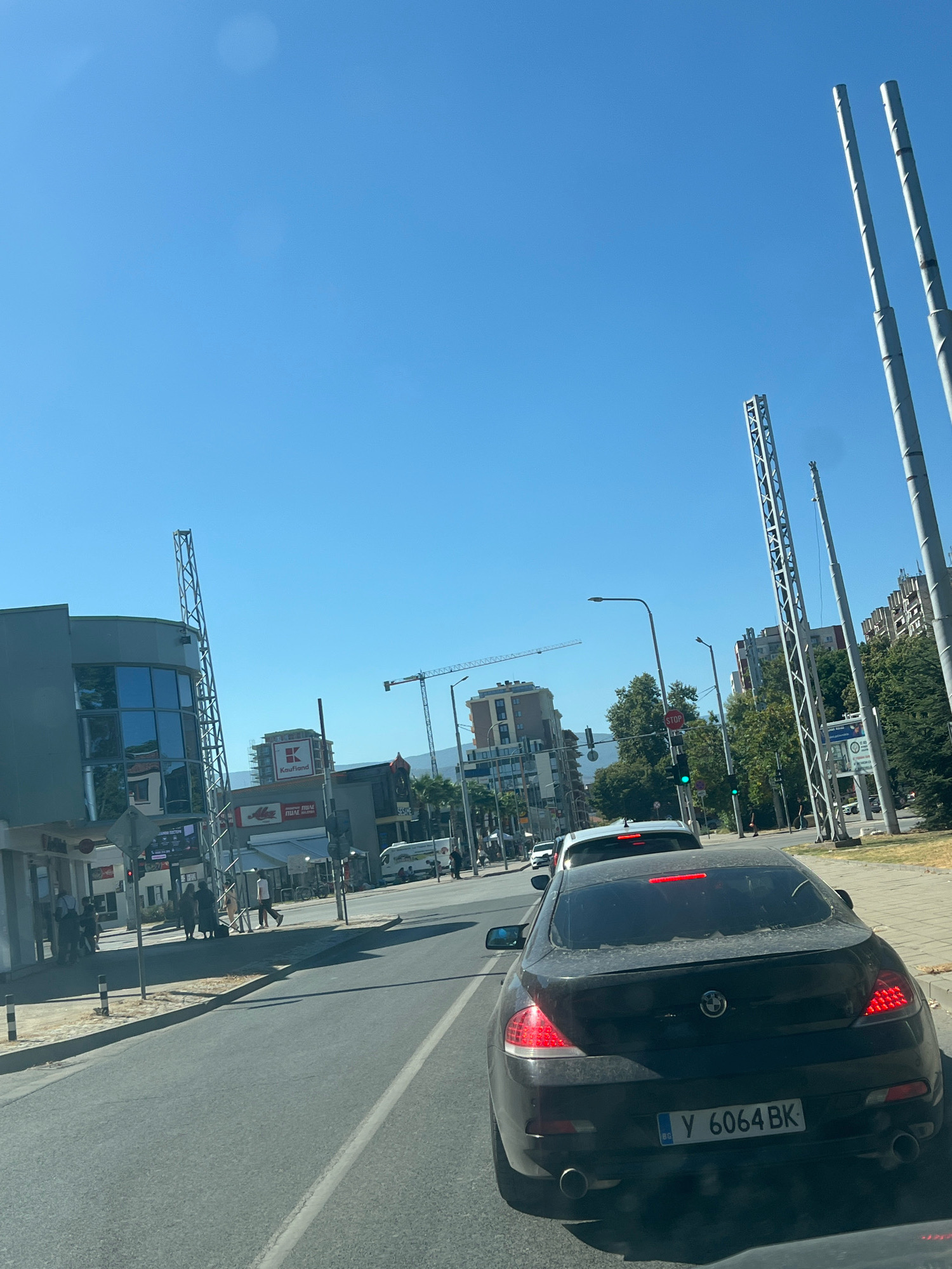 The image captures a street scene in Plovdiv, Bulgaria, on a clear, sunny day. In the foreground, a dark-colored BMW coupe with license plate "Y 6064 BK" is driving on a paved road. The car is dusty, indicating it has been driven. On the left side of the road, a modern building with large glass windows stands beside a metal tower structure. Further down the street, signs for "Kaufland" and "Alco" are visible, suggesting a commercial area. A construction crane is also present in the background, indicating ongoing development. Several pedestrians are walking on the sidewalk, and other vehicles, including a white van and a white sedan, are on the road. Traffic lights are visible, with one showing a green light. Tall, slender poles, possibly for lighting or telecommunication, line the right side of the road. The overall environment appears to be a bustling urban setting.