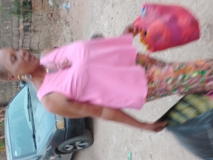 The image displays a female subject walking on an unpaved dirt and gravel path in Orie Uratta, Nigeria. The woman is in motion, wearing a pink sleeveless top, multi-colored patterned trousers, large gold-toned rectangular earrings, and a pendant. She carries a dark-colored bag in her left hand and a red plastic container in her right hand. In the background, a dark-colored sedan with five-spoke wheels is parked on the left, while a weathered concrete block wall is visible on the right.
