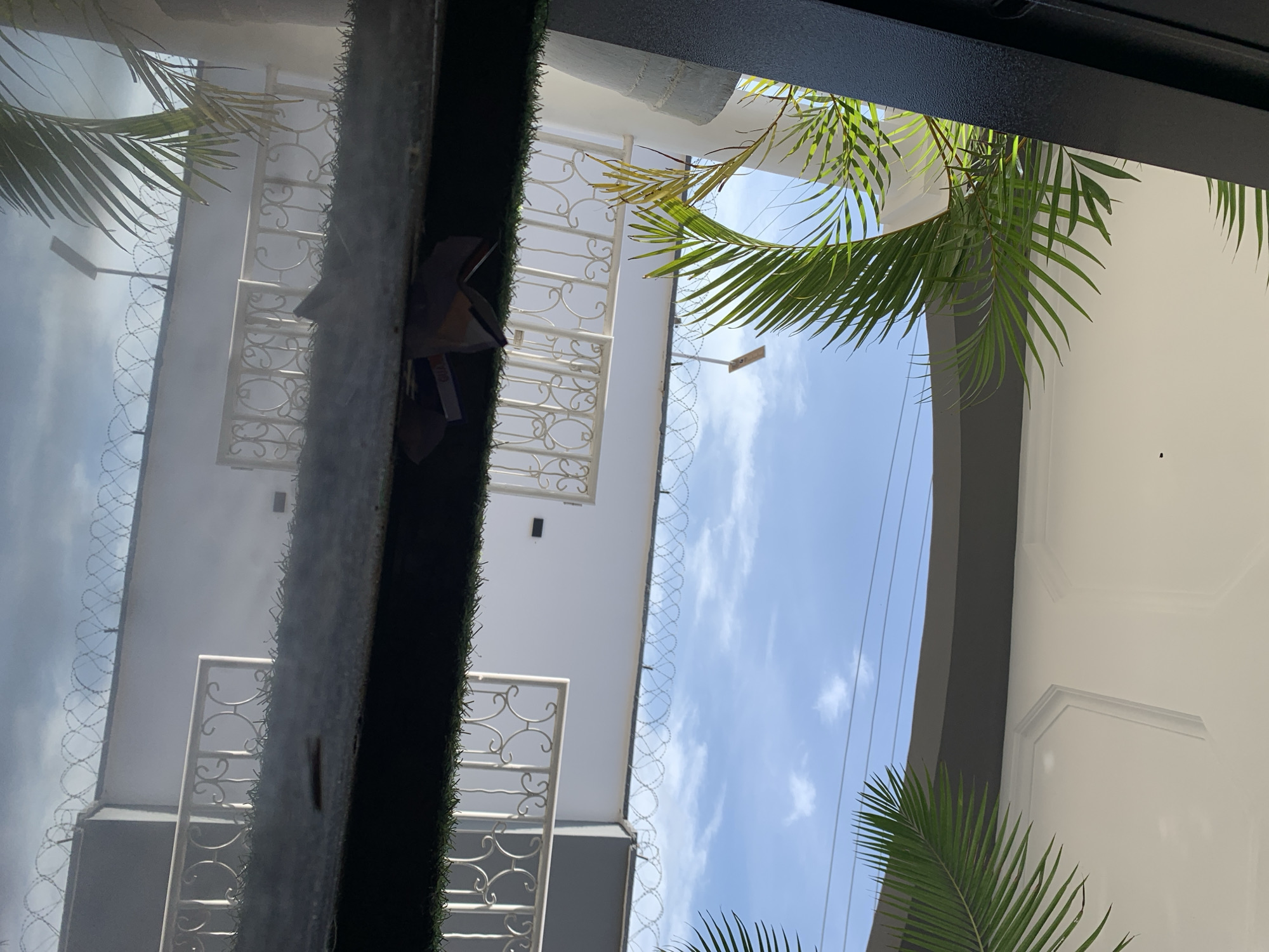 The image depicts an outdoor scene, possibly in Gura Top, Nigeria. A dark pillar is on the left side, partially obscuring a white arched ceiling with decorative molding. Green palm fronds are visible against the white ceiling and a blue sky with scattered clouds. Below, a white wall is topped with razor wire. A white decorative railing with scrolling patterns runs along the top of the wall. In the foreground, a reflective surface shows the scene upside down, with some artificial grass and a discarded paper object. A solar-powered light fixture is visible on the wall behind the razor wire. The overall impression is one of a residential or commercial property with security measures in place.
