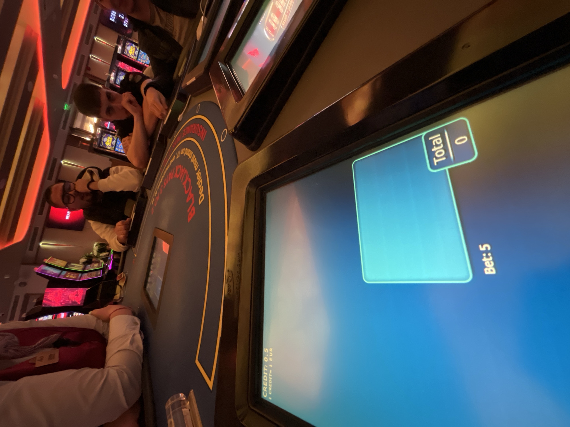 The image shows a low-angle view inside what appears to be a casino in Burgas, Bulgaria. Several slot machines with colorful displays are visible in the background. Two men are observed leaning on the edge of a Blackjack machine. The machine displays a blue screen with the text "Bet: 5" and "Total 0". The signage on the blackjack table indicates "Blackjack". In the foreground, part of another person's arm and white sleeve are visible, resting on the edge of the table. A plastic cup sits on the table. The lighting in the casino is a mix of red, orange, and blue.