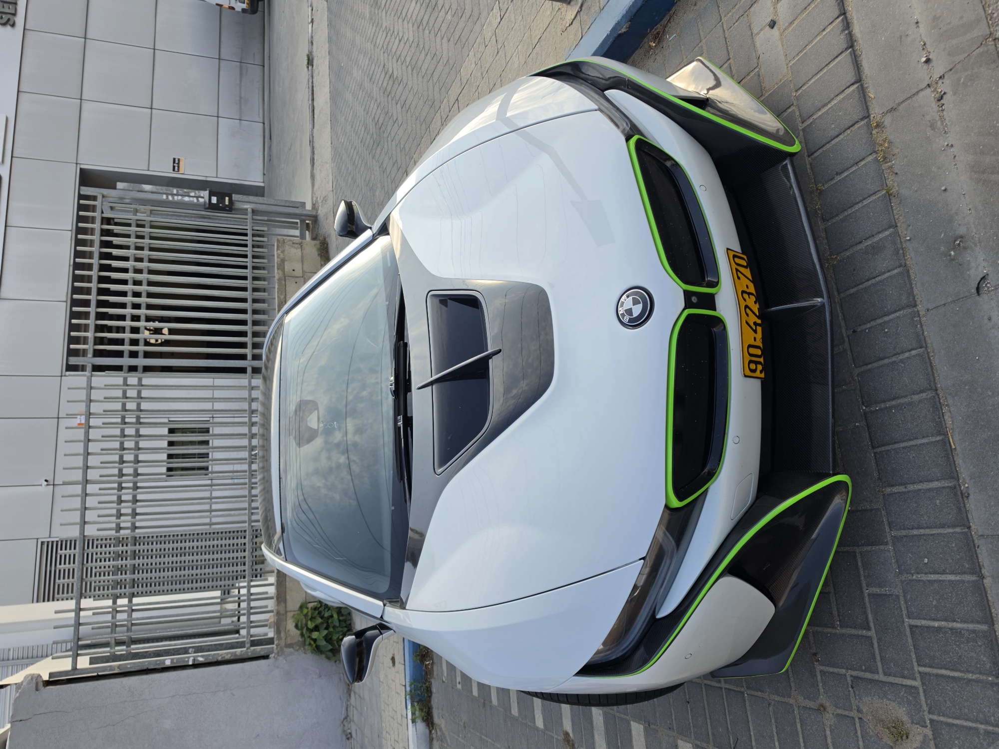 A white BMW i8 with green accents is parked on a brick sidewalk next to a modern building with a metal gate. The license plate reads "90-423-70," indicating it is in Rishon LeZion, Israel.