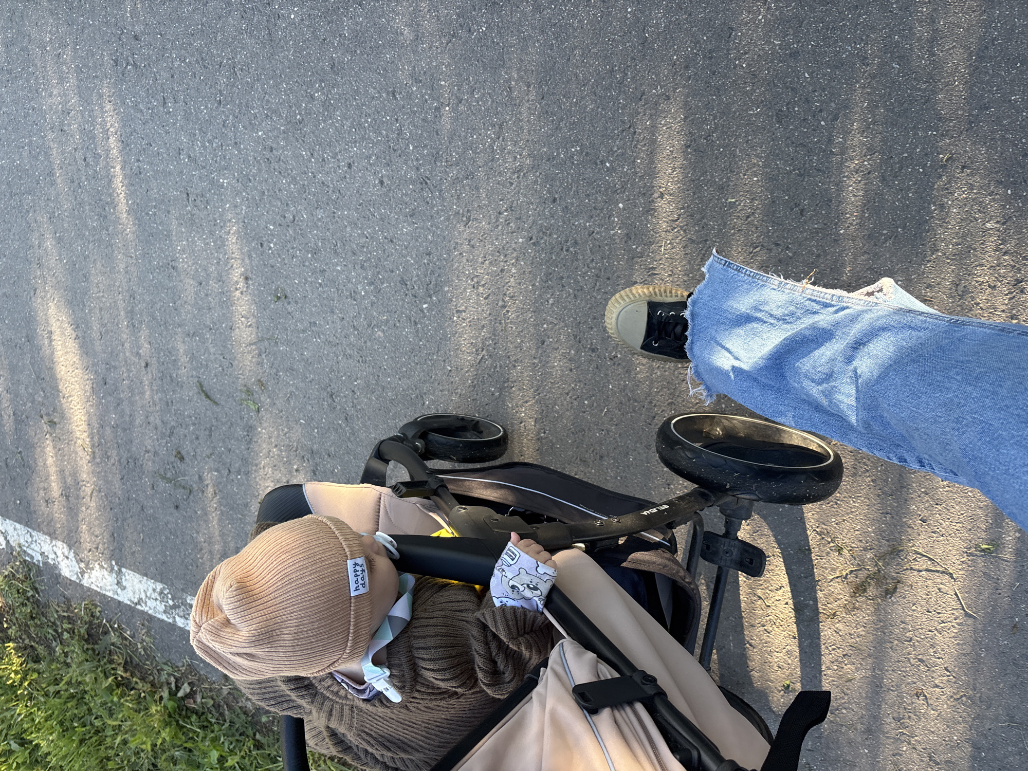 In the city of Мещёры, Russia, an outdoor scene depicts a close-up perspective of a person walking alongside a baby in a stroller. The pavement, marked by a white line on the left side, suggests a path or sidewalk. Sunlight casts dappled shadows across the asphalt. The baby, wearing a tan beanie with "happy days" embroidered on it, is nestled in a stroller with beige and brown fabric. The baby's hand, adorned with a patterned cloth, rests on the stroller's handle. The walker's legs, clad in ripped denim jeans, and a sneaker are visible in the lower right corner, indicating forward motion. The environment is characterized by a grassy verge on the left, suggesting a park or a tree-lined street.