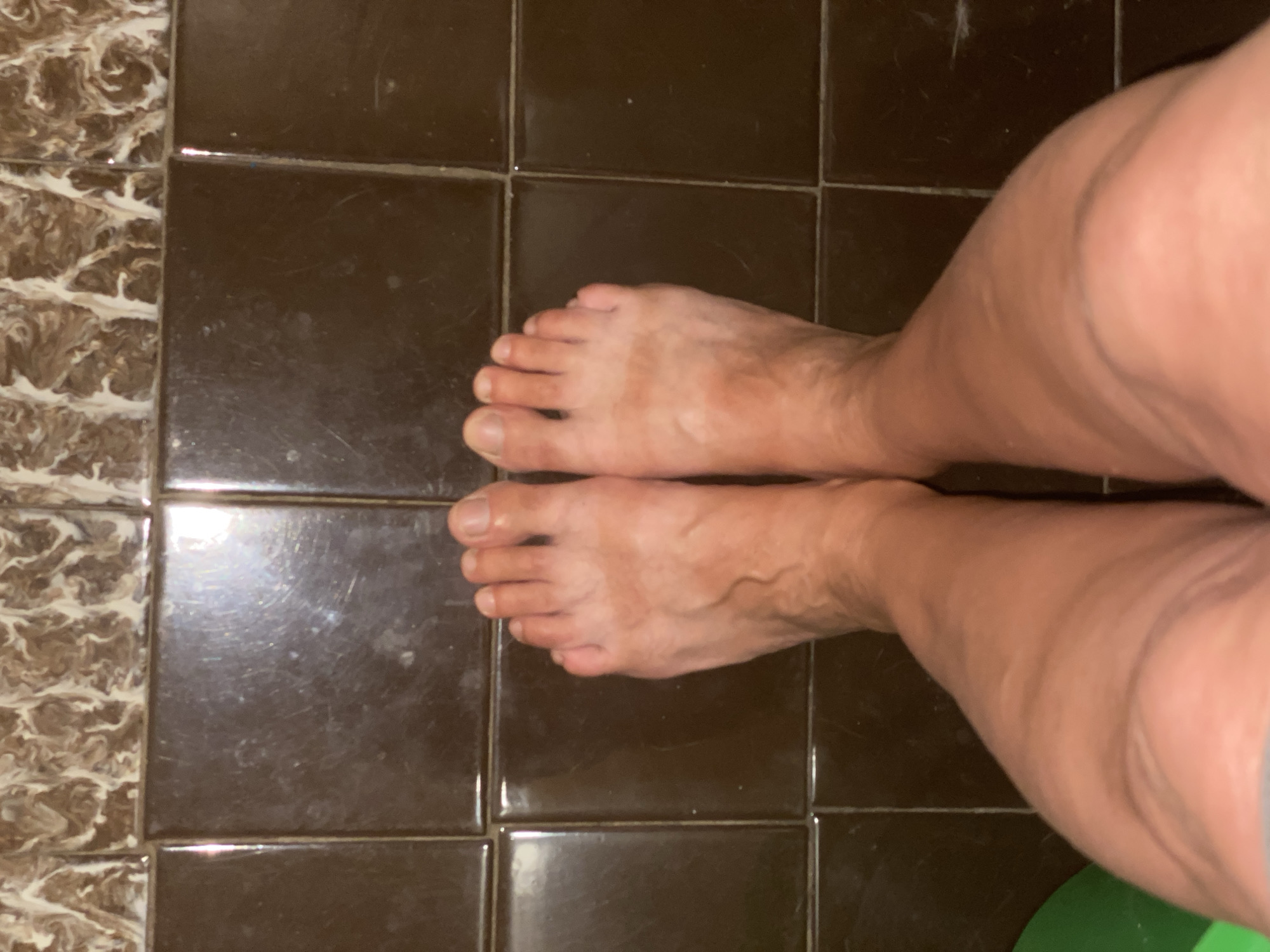 The image shows a close-up, overhead view of a person's bare feet and lower legs standing on a tiled floor. The tiles are dark brown and appear to be square. Above the brown tiles, there are tiles with a marbled pattern in brown and white. There are no identifiable landmarks or contextual clues within the image to determine its location in Novokuznetsk, Russia. The focus is entirely on the feet and lower legs, with no other subjects or objects clearly visible. The lighting suggests an indoor setting, possibly a bathroom or shower area due to the tiling.
