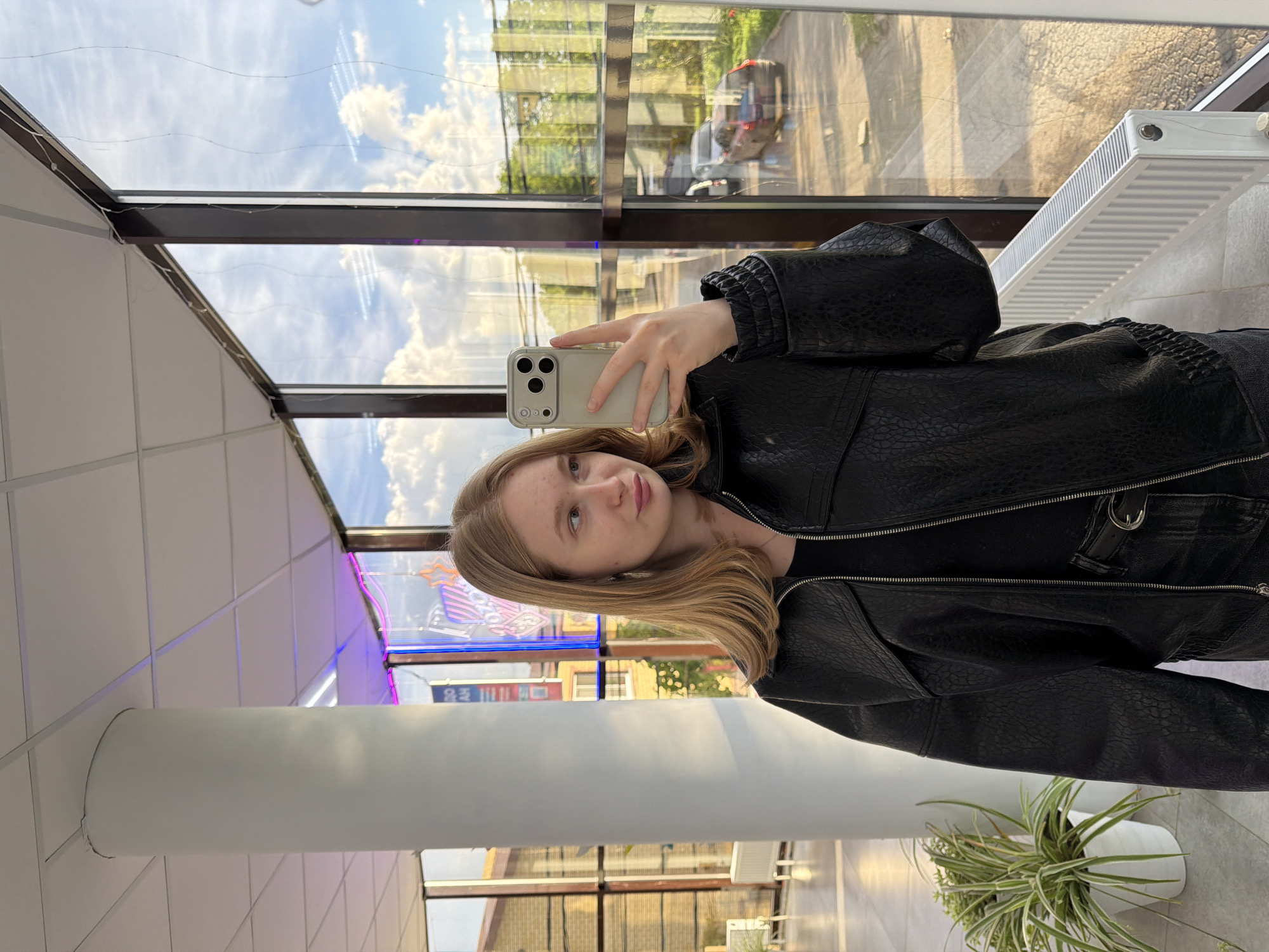 A fair-skinned young woman with blonde hair is taking a selfie with her iPhone inside a building in Rossiyskiy, Russia. She is wearing a black leather jacket. The building has large windows showing reflections of other buildings, the sky, and clouds. A white radiator is visible near the window. A large, white cylindrical support column is in the foreground. A potted plant is in the lower right corner. A store with neon lighting can be seen through the windows.