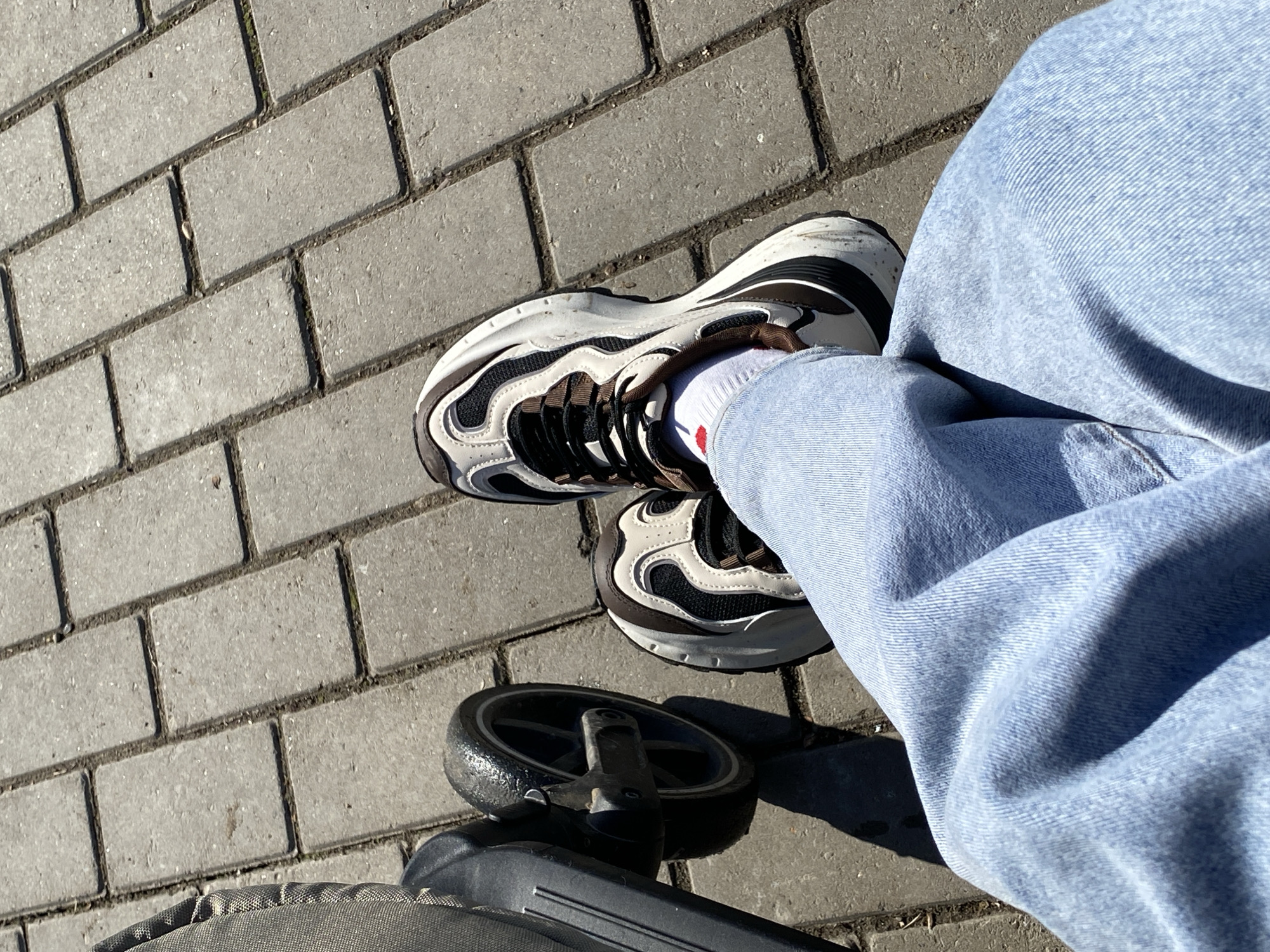 The image displays a low-angle, downward perspective of an individual's lower body and a partial view of a baby stroller, situated on a paved surface under bright sunlight in Chaikovsky, Russia. The individual is wearing light blue denim trousers and white ankle-length socks, the left of which features a small red embroidered heart motif. Both feet are clad in 'chunky' athletic sneakers, characterized by a design incorporating white, black mesh, and brown accents, black laces, and prominent multi-layered white and black soles. Minor scuff marks are visible on the white sections of the soles. To the immediate left, a black-rimmed stroller wheel with a rubber tire is visible, along with a section of the stroller's dark grey fabric and black plastic frame. The ground surface is composed of light grey rectangular concrete pavers laid in a stretcher bond pattern, with discernible grout lines that contain minor debris and organic matter. Distinct shadows are cast across the pavers, indicating strong overhead sunlight.