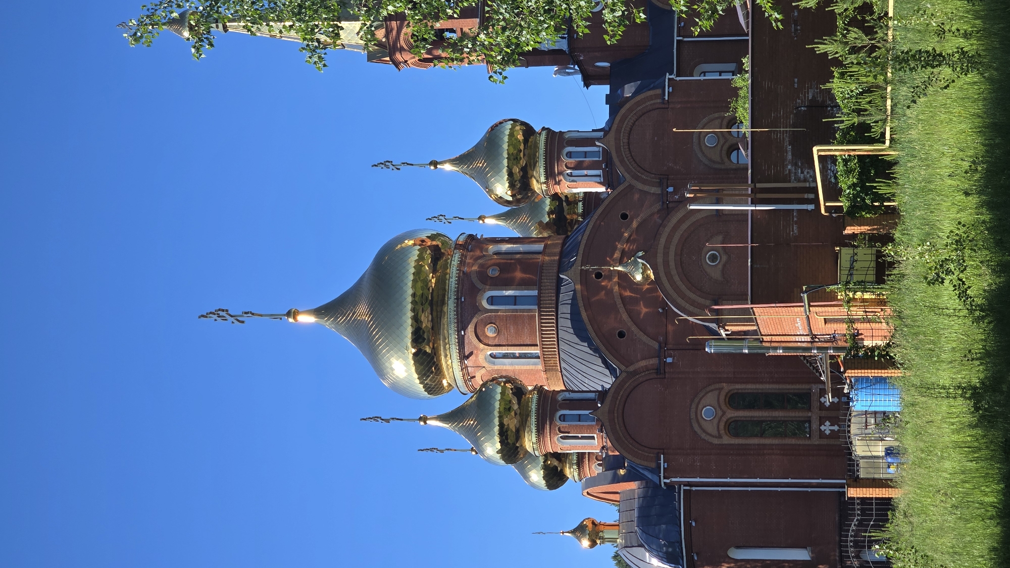 The image shows the Church of the Transfiguration in Chaikovsky, Russia. The church is a large, red brick building with multiple golden domes, each topped with a cross. The sky is clear and blue. In the foreground, there is a grassy field with some trees and a fence. There are also some industrial elements visible, such as pipes and a blue tank, which are part of the church's infrastructure. The sun is shining brightly, casting shadows on the building and the surrounding area.