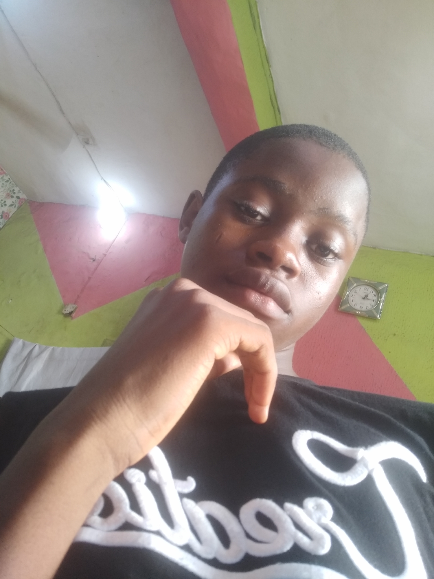 The image is a selfie taken from a low angle looking up at a young person with dark skin and short hair. They are wearing a black t-shirt with white lettering that is partially visible. The person has their right hand resting on their chin, with their fingers extended. The background appears to be a room with a light-colored ceiling and walls painted in geometric patterns of pink and green. A small, square clock with a white face and black hands is visible on the wall to the right. A light fixture is also visible on the ceiling, emitting a bright light. The overall impression is of an indoor environment, possibly a home. There is no information within the image to suggest a specific location within Shangisha, Nigeria.