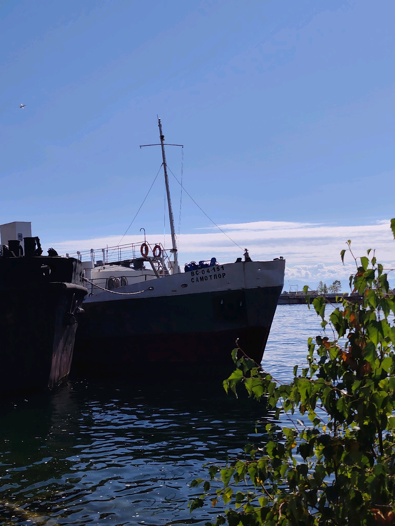 An image of two ships docked in the water near the city of Baikal, Russia. The ship in the foreground on the left is dark and partially obscured. The ship on the right is white with a red waterline and has the Cyrillic text "ВС-04-151 САМОТЛОР" on its bow. A mast with rigging extends from the ship on the right, and two life preservers are visible on its deck railing. The water is blue with ripples, and a patch of green foliage is in the lower right corner of the frame. The sky is bright blue with scattered clouds, and a small white airplane is visible in the upper left corner.