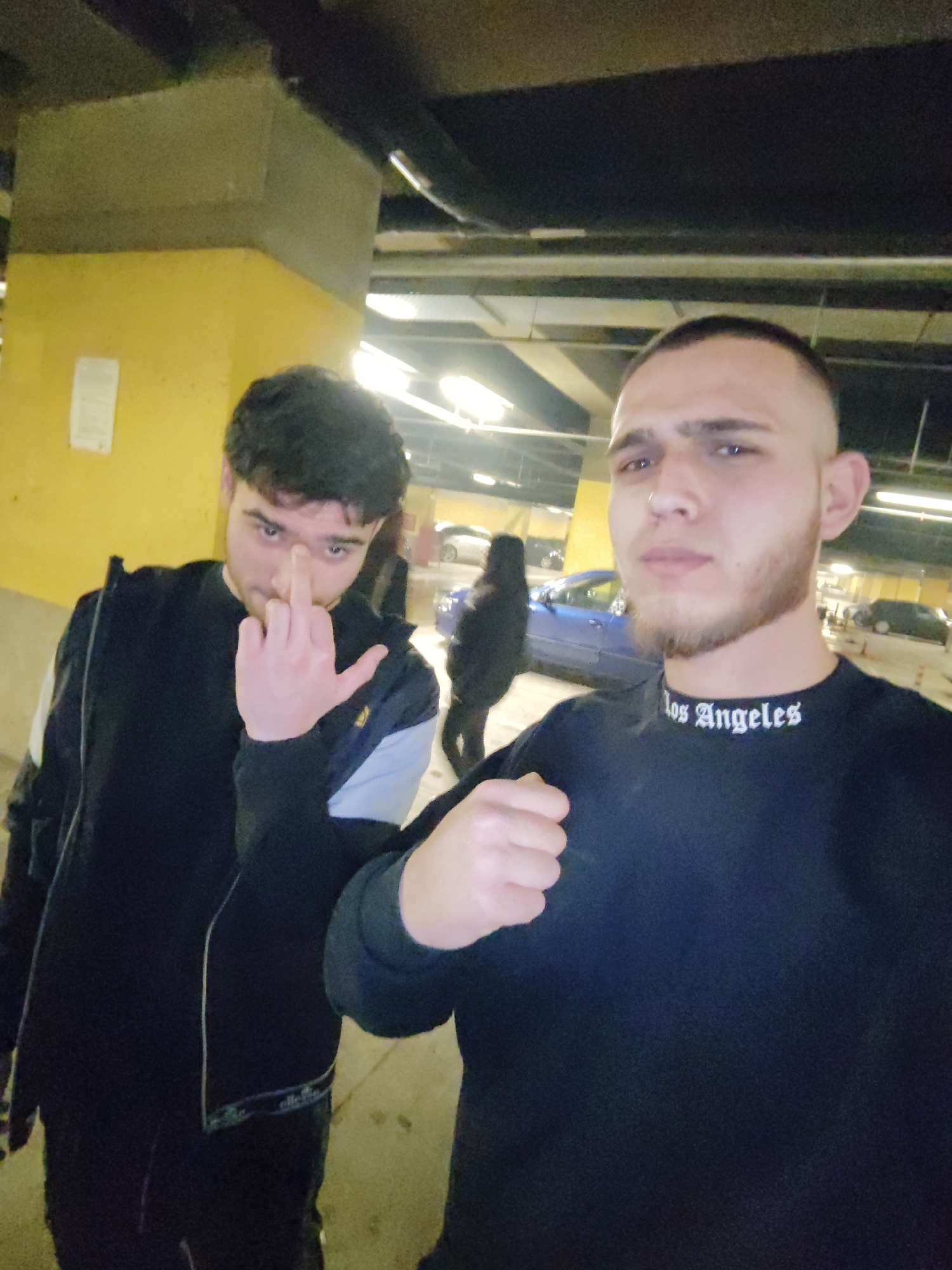 Two young men are the primary subjects of this medium shot, likely taken in an underground parking garage. The man on the right, closer to the camera, wears a black long-sleeved shirt with "Los Angeles" printed in white around the neck. He has a close-cropped haircut and a light beard. His right hand is clenched in a fist. The man on the left, standing slightly behind, wears a dark jacket with white and light blue details. He is holding up his middle finger.
In the background, partially obscured, are parked cars, a person walking, and a yellow support column. The overhead lighting is visible, suggesting an indoor environment. The location is likely Sofia, Bulgaria. The overall atmosphere of the photograph is casual and potentially provocative.