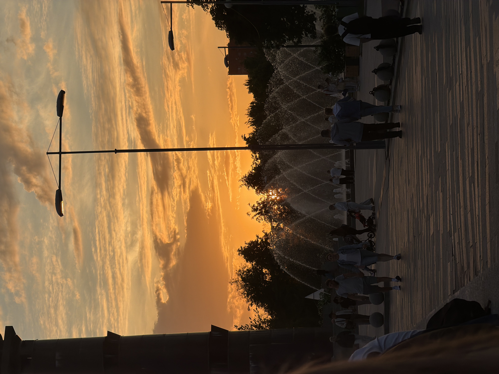This image captures a vibrant sunset over a public square or park in Saint Petersburg, Russia. The sky dominates the upper portion of the frame, painted with warm, glowing hues of orange, yellow, and soft blue, featuring long, streaky clouds illuminated by the setting sun.
The low sun, a bright golden orb, is visible just above a dense line of trees that forms a dark silhouette across the middle ground. In front of the trees, a large fountain sprays multiple jets of water, which catch the golden light and create a glittering, dynamic display.
Numerous people are present in the scene, mostly silhouetted against the bright sky and illuminated water. They are scattered across a paved area that stretches from the foreground to the mid-ground, showing long, distinct shadows cast by the low sun. Individuals appear to be enjoying an evening stroll or gathering in the area; one person is pushing a baby stroller, while others walk or stand.
Several modern-looking lampposts, with sleek, elongated light fixtures, are prominently silhouetted against the colorful sky. One notable lamppost features two horizontal arms. In the background, behind the trees and fountain, a dark, blocky building is faintly visible. Dark, spherical objects, possibly decorative bollards or seating, are placed along the paved path. The overall mood is peaceful and enjoyable, capturing a beautiful golden hour moment in a lively urban setting. No visible text is present in the image.