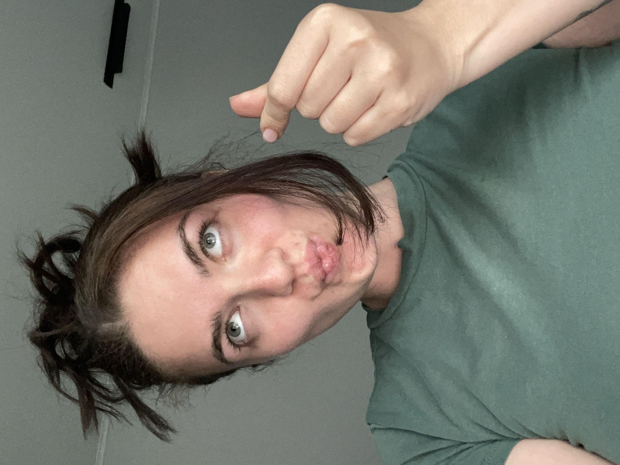 A woman is facing the camera with her head tilted to the left. She has brown hair pulled up in a messy bun and is wearing a green t-shirt. Her lips are pursed in a kiss. She is holding up her hand above her head, her fingers in a pinching gesture. The background is a plain gray wall with a black object hanging near the top left corner. The photo was taken indoors. The location, Мосметаллоконструкция Онт, Russia, is not discernible from the image.