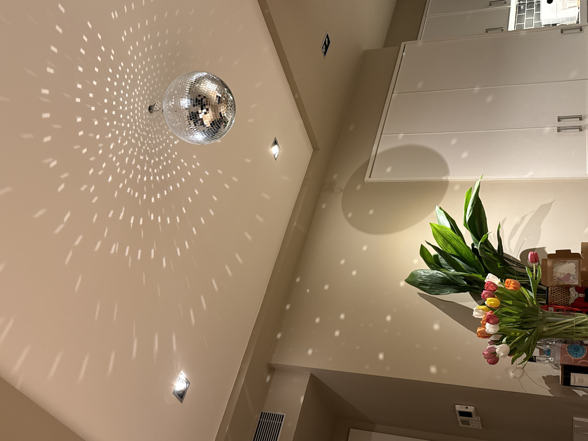 The image depicts an interior space, possibly a modern apartment. A mirrored disco ball hangs from the ceiling, reflecting light onto the walls and other surfaces in a pattern of small, rectangular shapes. The walls are painted in a neutral, light beige color. Recessed lighting fixtures are visible on the ceiling. A bouquet of tulips and large leaves is placed on a countertop. Above the flowers, white cabinetry with horizontal handles is seen. The location seems to be indoors, possibly a residence. There is no evidence in the image to support that this is Barcelona, Spain.