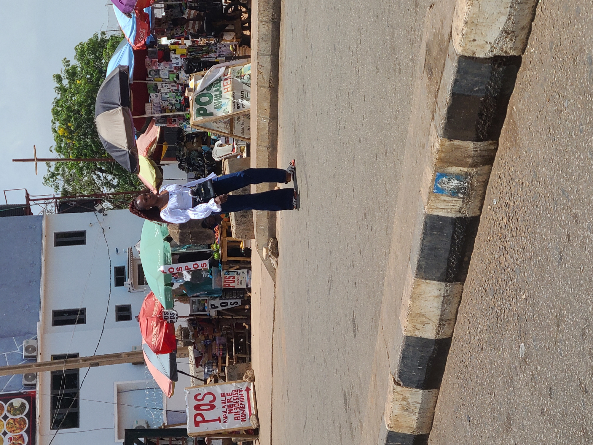 The image depicts a street scene in Jos, Nigeria. A woman with dark hair pulled back, wearing a white blouse and dark pants, stands on the sidewalk. Behind her are vendor stalls covered by various umbrellas, including a Coca-Cola branded one. Signs advertising "POS AVAILABLE HERE" are visible in multiple locations. The street is paved, and a black and white striped curb separates it from the sidewalk. A multi-story building is visible in the background.