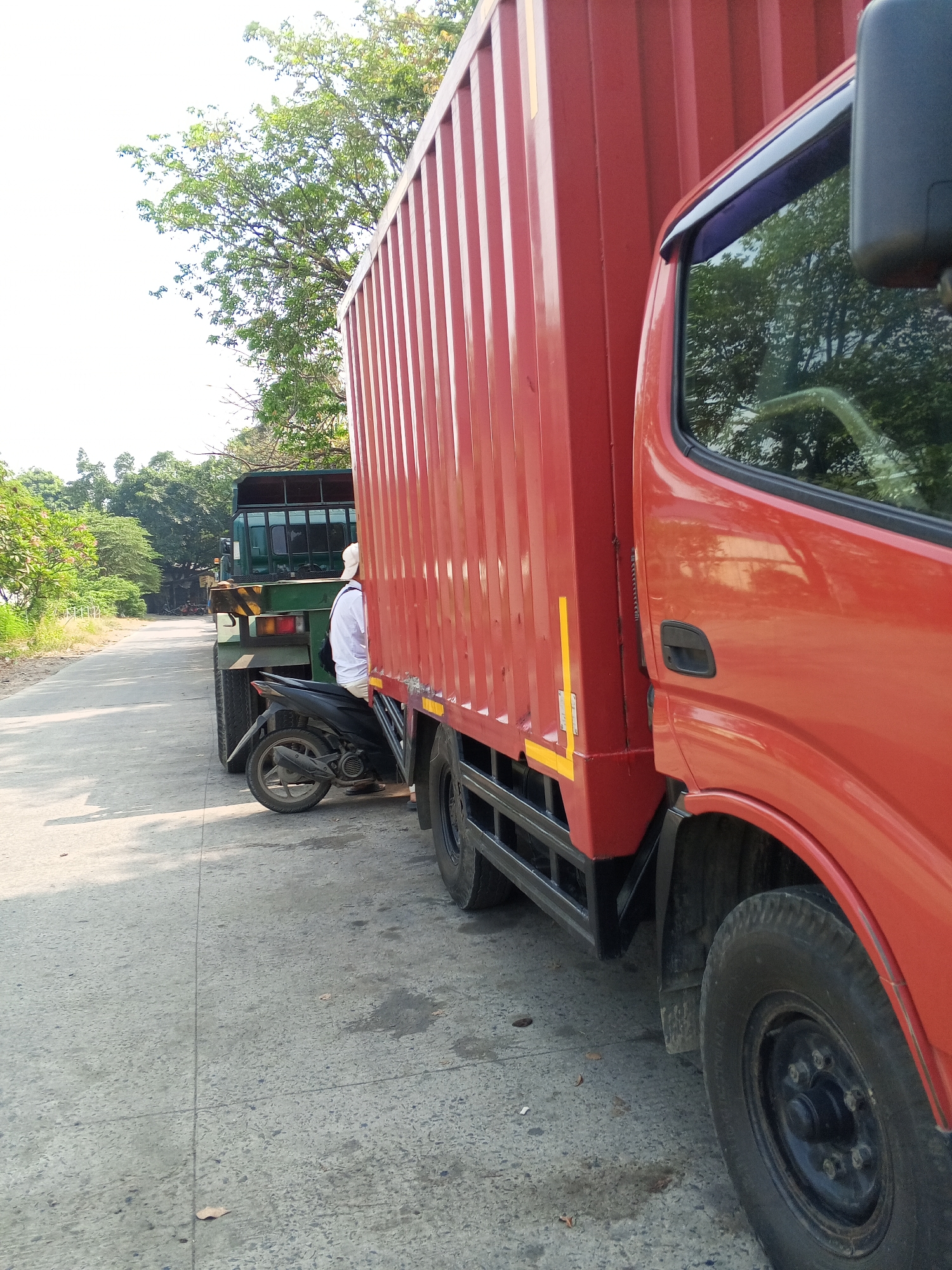 A person wearing a white shirt and a white cap is seated on a black motorcycle, parked alongside a red truck. The motorcycle is positioned between the red truck and a green truck. The scene appears to be on a paved road with trees lining the left side. The sky is bright and clear. The image is taken from a low angle, focusing on the side of the red truck.