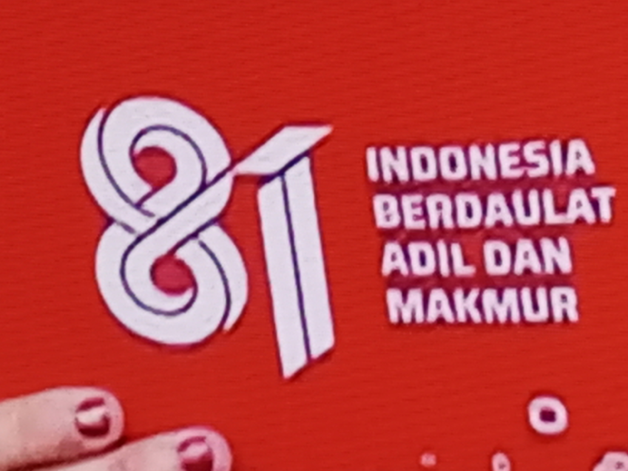 The image shows a red background with white text and graphic elements. On the left, there is a large white graphic that appears to be the number "81", stylized with loops and a sharp vertical line. To the right of the "81" graphic, there is text arranged in three lines. The first line reads "INDONESIA", the second line reads "BERDAULAT", and the third line reads "ADIL DAN MAKMUR". Below the text, there are several small white circular and oval shapes. In the lower left corner, there are two fingers with dark red nail polish. The image appears to be a photograph or a screen capture of a banner, sign, or digital display. There is no information in the image that indicates a specific location in the city of Banyudono, Indonesia.