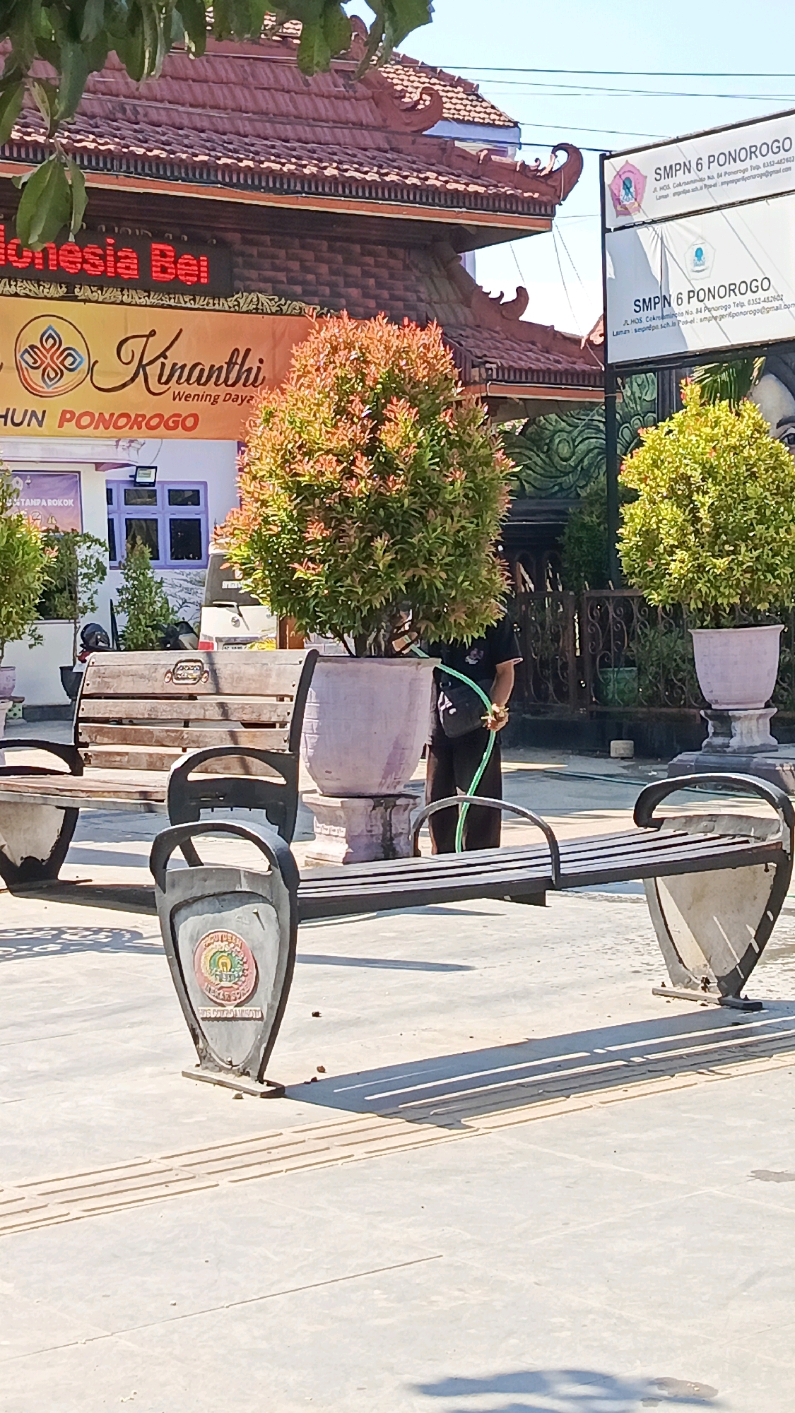 The image depicts a scene in front of SMPN 6 Ponorogo in Ponorogo, Indonesia. A person is watering a tree in a large pot with a green hose. Several park benches are visible in the foreground, some made of wood and others with metal frames. A sign for SMPN 6 Ponorogo is displayed on the right side of the image. The environment appears to be an outdoor public space with a paved ground and some landscaping. The sky is clear and blue, indicating a sunny day.