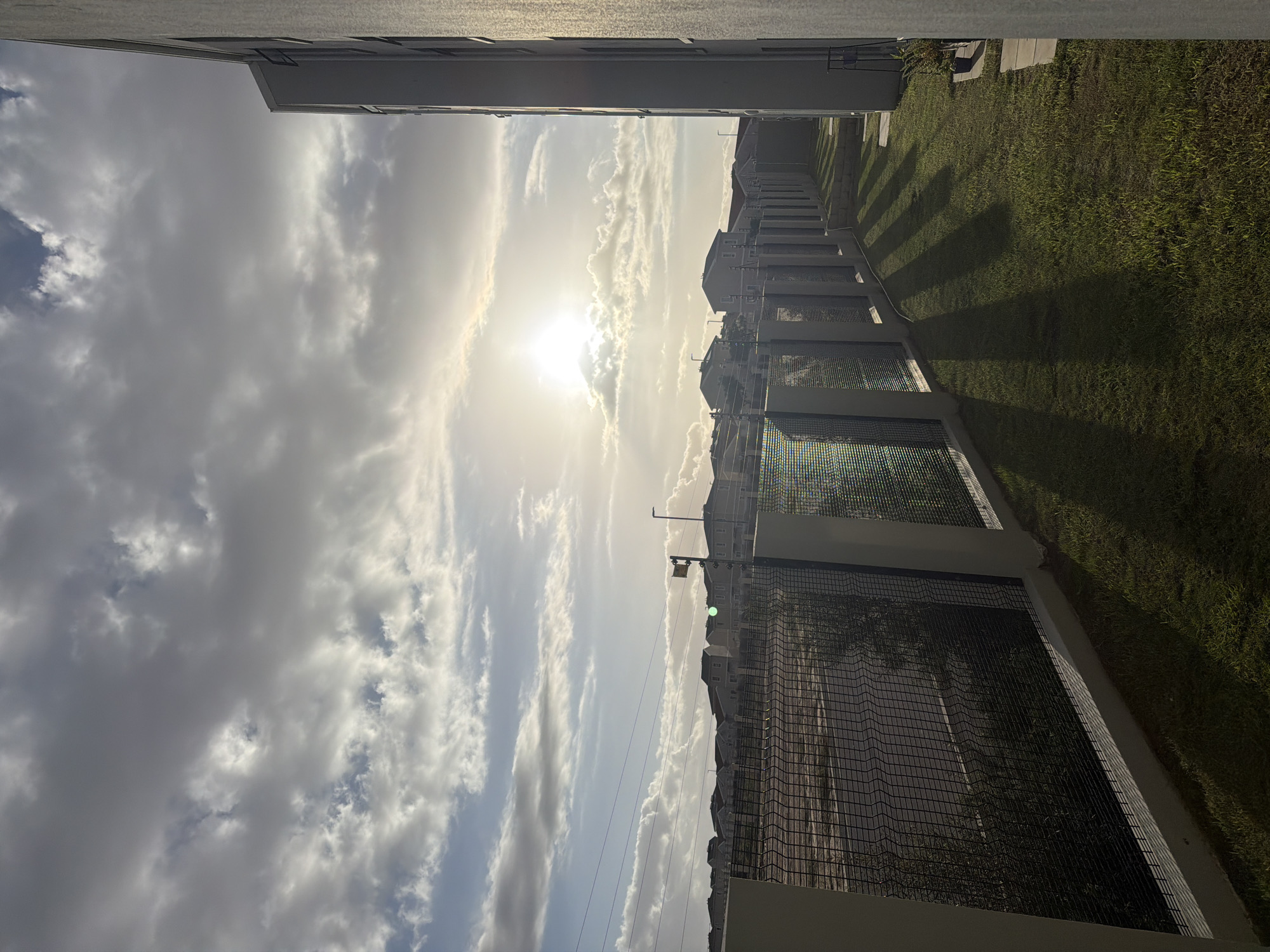 The image shows a view from what appears to be a high vantage point, likely a building. The sky dominates the scene with a mix of bright sun and clouds. Below the sky are buildings and a fence line running parallel to the buildings. Adjacent to the fence line is a green grassy area. The image is taken from what may be a balcony in Cape Town, South Africa. There are no people visible in the image. The architectural style of the buildings is modern. The presence of the fence suggests a boundary between the buildings and the grassy area. Shadows cast on the grass indicate a time of day when the sun is at an angle.