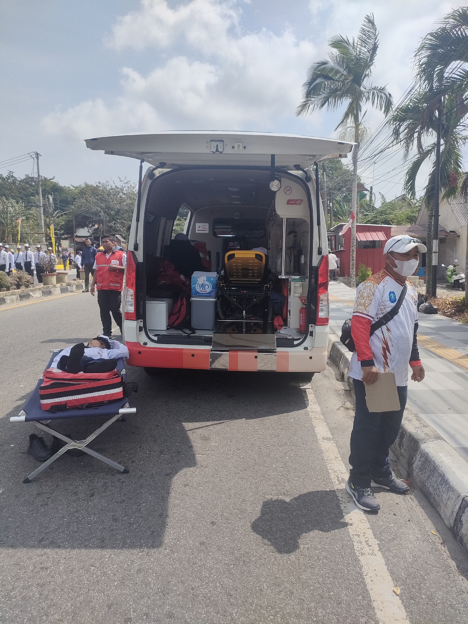 The image depicts an outdoor scene in Sungailiat, Indonesia, featuring an open ambulance with its rear doors extended upwards. A person in a white shirt, dark pants, and a white cap with a face mask is standing on the right side of the image, holding a piece of paper. To the left of the ambulance, a portable cot is set up on the asphalt road, with a person lying on it, wearing a light-colored uniform and dark pants. Behind the cot, another person wearing a red vest is visible, facing towards the ambulance. The background shows a street lined with trees, including palm trees, buildings, and a group of people in the distance who appear to be in uniform. The sky is partly cloudy. The scene suggests a medical or emergency response situation in a public area.