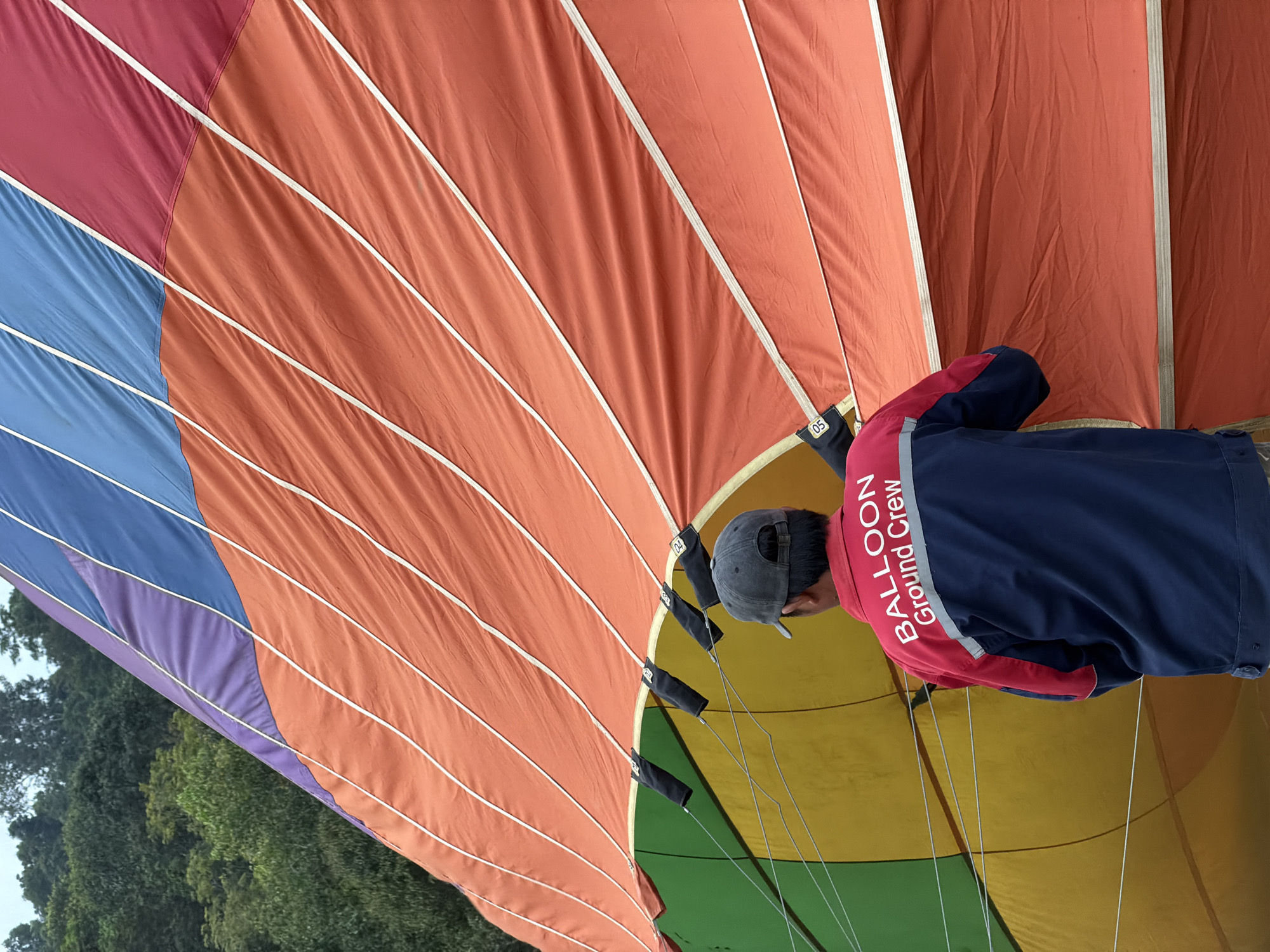 The media file captures a close-up view of a hot air balloon from the inside, with a person wearing a "BALLOON Ground Crew" jacket visible. The balloon's fabric is composed of large, colorful panels in shades of red, orange, blue, purple, yellow, and green, separated by white seams. The person, seen from behind, is wearing a gray baseball cap and a blue and red jacket with white text that reads "BALLOON" and "Ground Crew". They appear to be attending to the balloon's rigging, with dark fabric loops and suspension lines visible. The scene is set outdoors, with hints of green foliage and a cloudy sky visible in the upper left corner, suggesting it might be an overcast day. The overall impression is one of preparation for a hot air balloon flight, with the ground crew member engaged in their duties. The location context provided is Vang Vieng, Laos.