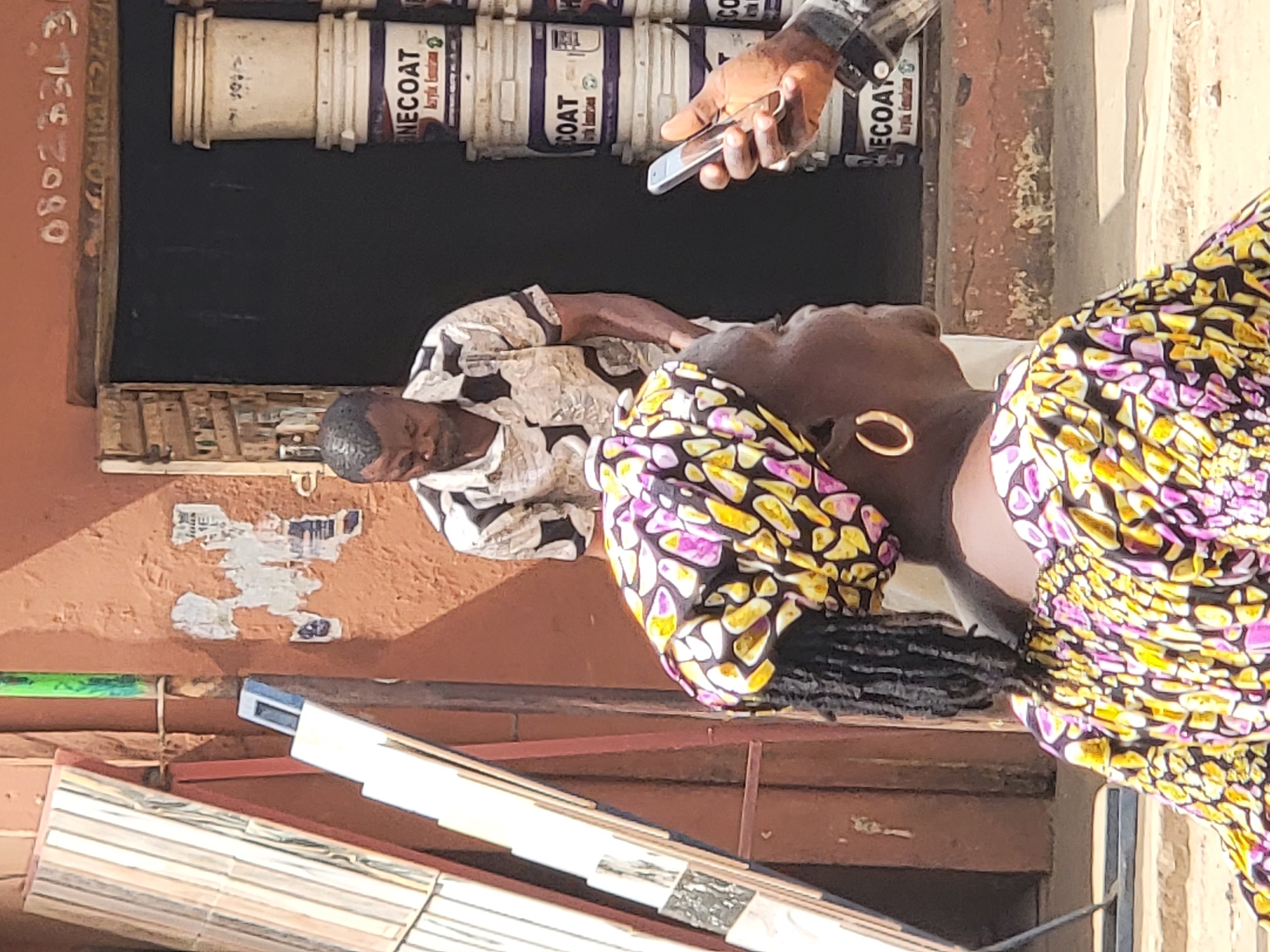 In Maiburiji, Nigeria, the image shows three individuals near a building displaying paint buckets labeled "ONE COAT." One man in a patterned shirt is seated. A second man holds a phone. A woman wearing a colorful headwrap and earrings stands nearby. Tiles lean against the building's exterior, which has a phone number painted on it.