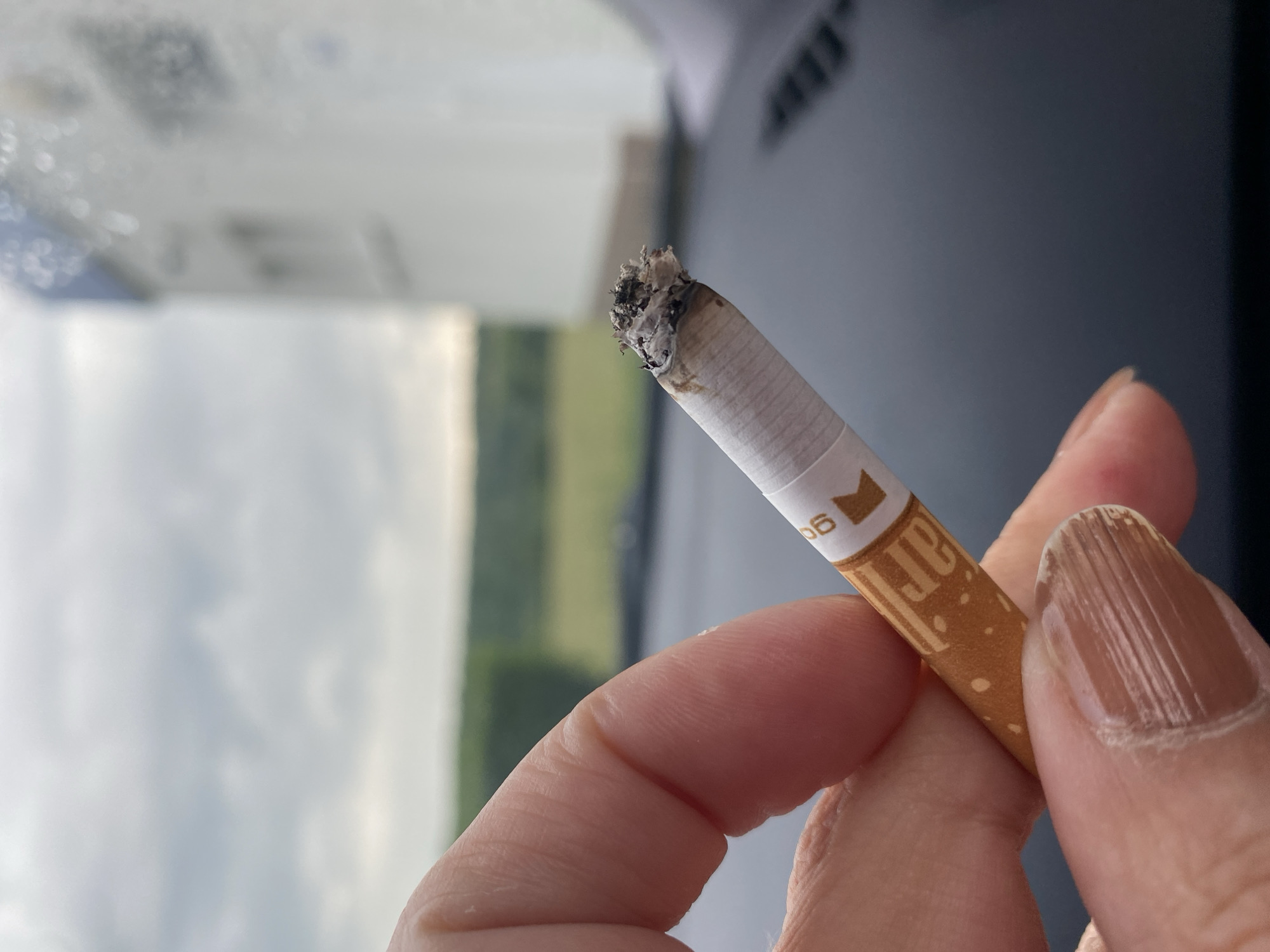 An extreme close-up shot shows a person's hand holding a lit cigarette. The cigarette is partially burned, with ash at the tip. The cigarette filter is white with an orange band and text that appears to be "ar" and "06". The background is blurred, showing a cloudy sky, green trees, and a white building. The overall impression is one of a casual moment outdoors. The location of Bílina, Czechia is not visually present in the image.