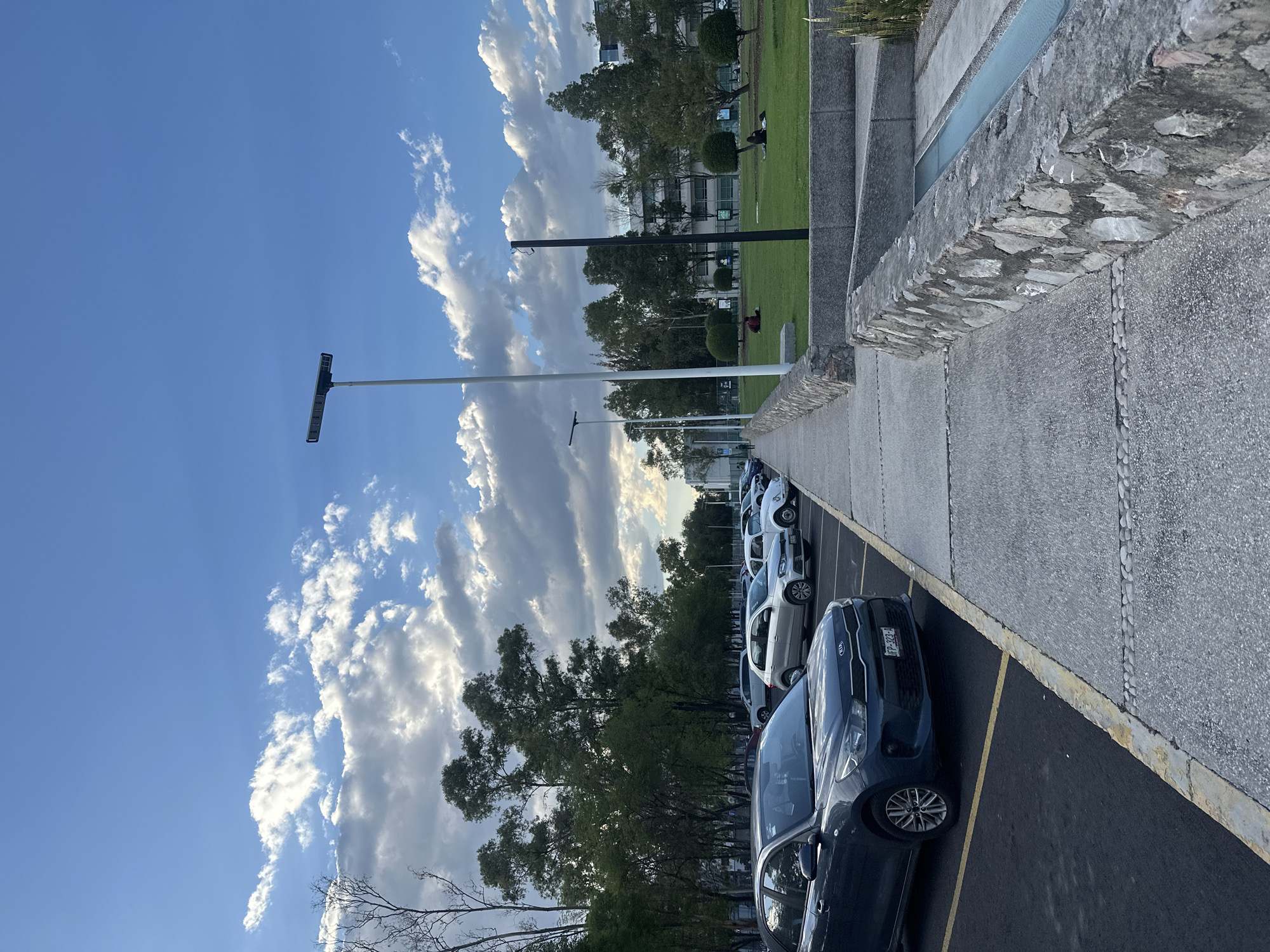 The image shows a parking lot in an urban environment. Several cars are parked in designated spaces marked by yellow lines. A low wall separates the parking lot from a grassy area with trees and a building in the background. Two people are sitting on the grass. The sky is blue with scattered white clouds, and there are tall lampposts along the street.