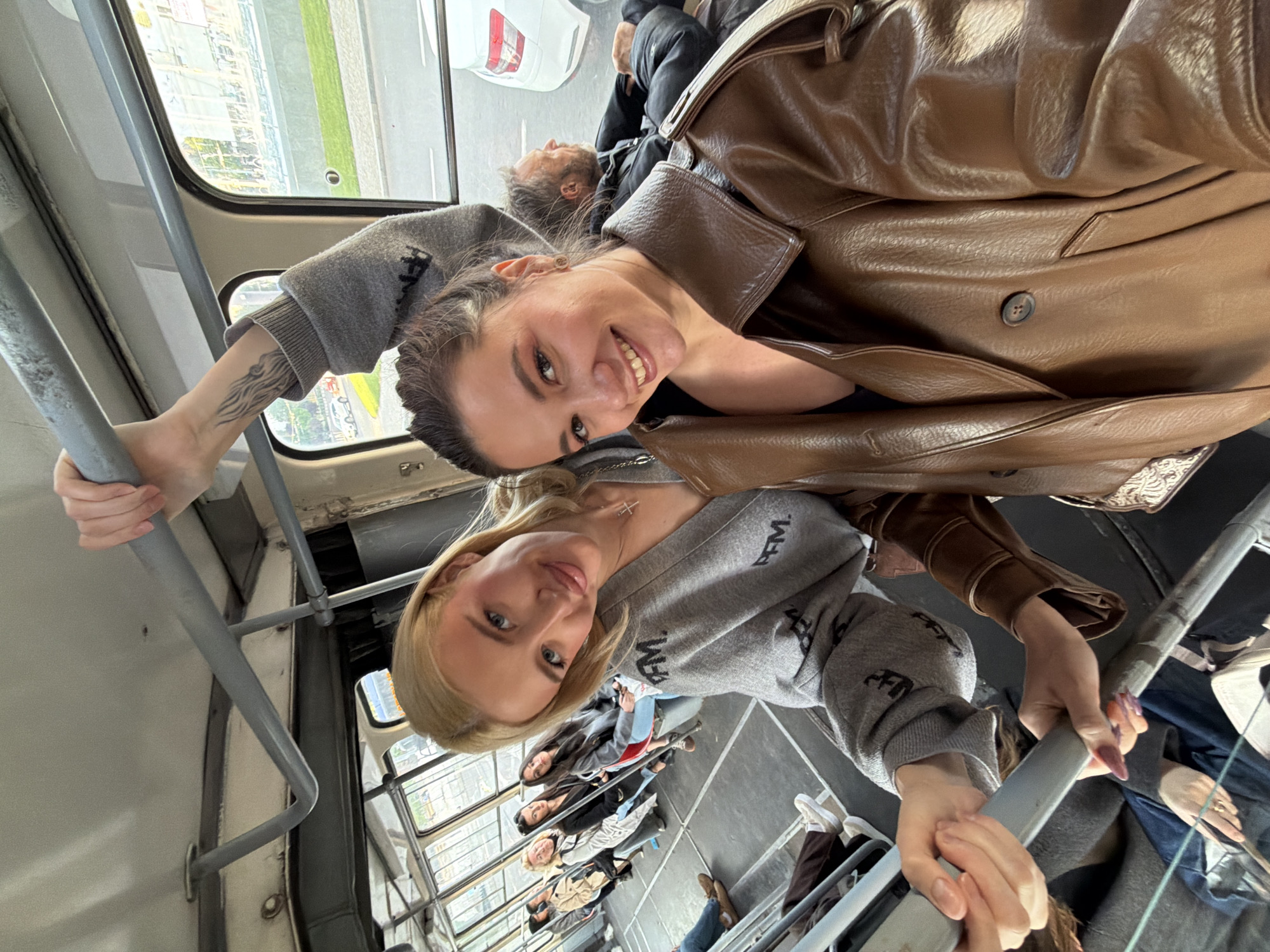 This candid photo captures two women taking a selfie inside a public transport vehicle, likely a tram or bus, in Belgrade, Serbia. The image is taken from a low angle, looking upwards at the subjects, creating a dynamic and informal feel.
In the foreground, a woman with dark hair and a brown leather jacket smiles broadly at the camera. She has a visible tattoo of a wavy, wing-like design on her left forearm. Beside her, a blonde woman in a grey sweatshirt also smiles, holding onto a metal grab bar with long, dark-painted nails. Both women's sweatshirts feature stylized black text that appears to be "FBM" or similar, repeatedly printed.
The setting is an older model public transport vehicle, characterized by its functional interior, including numerous grey metal grab bars and large windows. Through the windows, an urban landscape is visible, with buildings, a road, and green areas, indicating daytime. In the background, other passengers are present, including a man with a beard sitting down, and several blurry figures further back, all appearing to be riding the vehicle.
The overall mood is cheerful, conveyed by the smiles of the two primary subjects. The image captures a moment of everyday life and interaction within the context of public transportation.