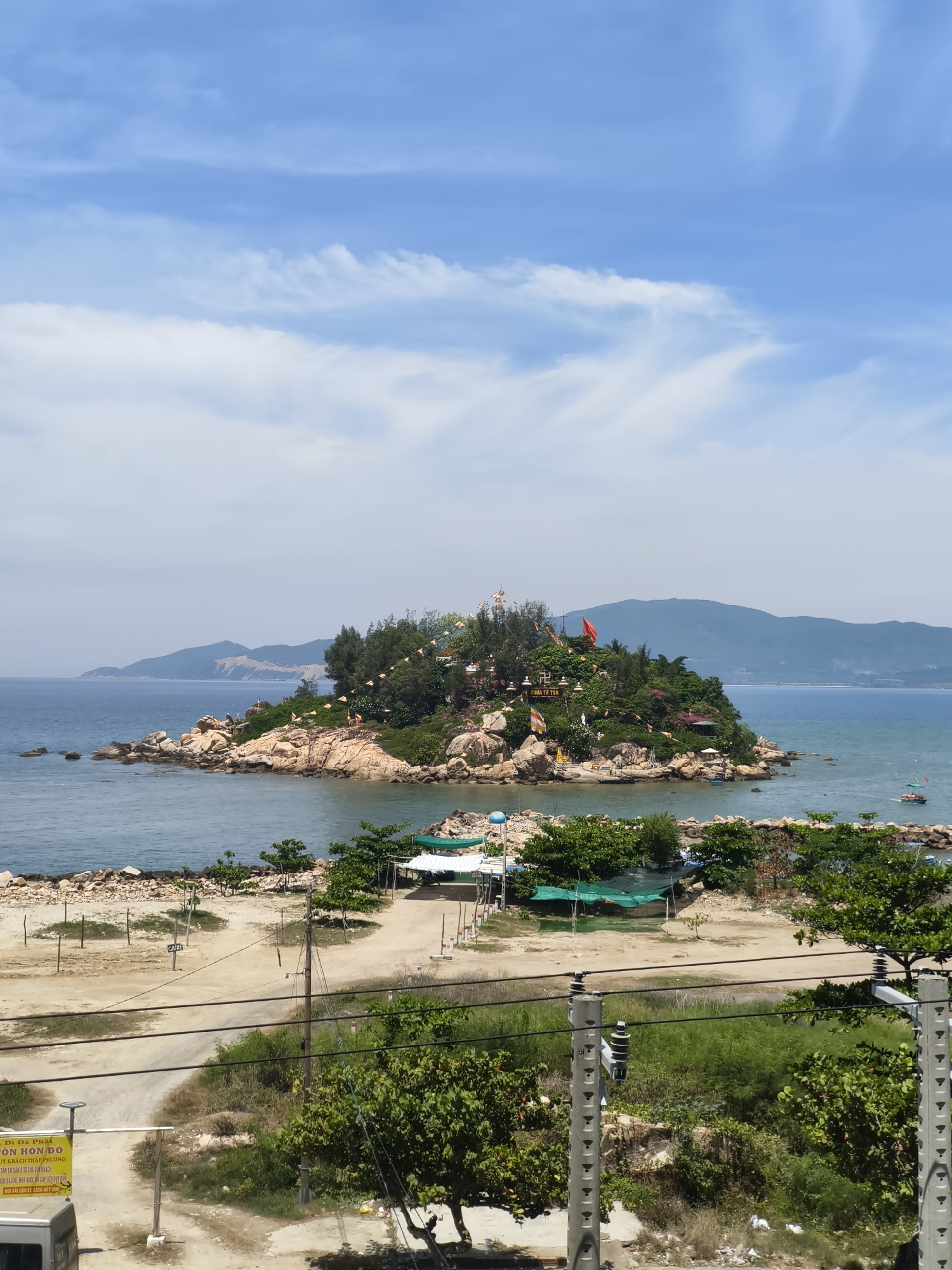 An elevated, vertical, full shot captures a scenic view of a small, rocky island adorned with vegetation and structures, jutting into the calm, blue sea under a partly cloudy sky. The island, which is a temple complex, is situated off the coast of Nha Trang, Vietnam, a popular tourist destination. The foreground shows a dusty, unpaved area with scattered trees, power lines, and a section of what appears to be a parking lot. A yellow sign with Vietnamese text is visible on the left side of the frame. In the midground, the island is teeming with greenery, rocky outcrops, and various religious icons and structures, including prayer flags and what seem to be statues. A red flag flies prominently on the island. The sea surrounding the island is a tranquil blue, with a small boat visible in the distance. The background features hazy, mountainous terrain receding into the horizon. The overall impression is one of a serene coastal landscape with a significant cultural and religious site.