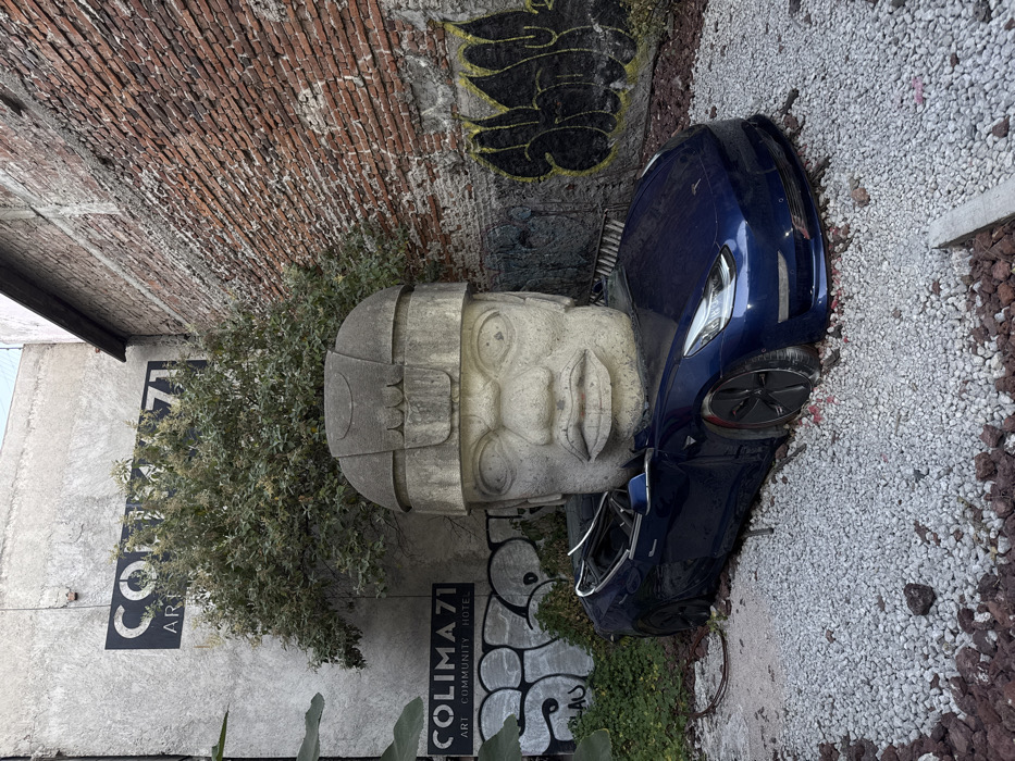 A blue Tesla rests beside a large stone Olmec head statue. The backdrop includes a weathered brick wall and a concrete wall with "COLIMA 71" signage. Gravel covers the ground where the car is parked. Some graffiti is visible on the wall near the Olmec head. Vegetation is interspersed around the statue and signage. The Tesla's open driver's side door hints at recent activity.