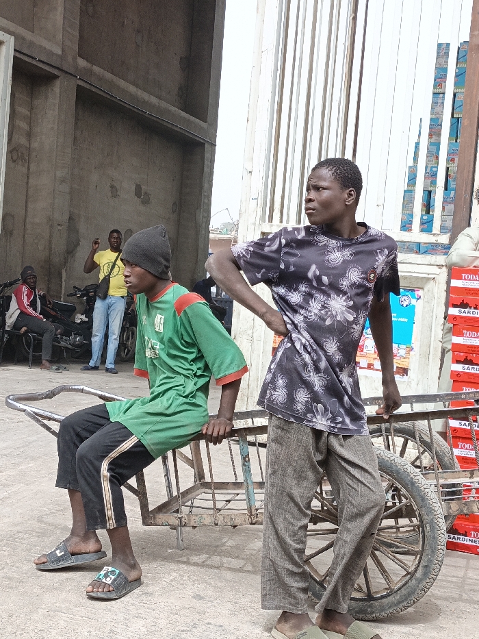 In Fagge C, Nigeria, an outdoor scene depicts five males and commercial activity under bright daylight. In the foreground, a young male with dark skin sits on a rusty, two-wheeled metal pushcart. He wears a dark grey beanie, a green t-shirt with red trim, black track pants, and black slide sandals, looking left. Standing to his right, another young male with dark skin wears a black t-shirt with white floral patterns, loose light brown pants, and light green slide sandals. He stands with his right hand on his hip, also looking left.
In the midground, to the left of the pushcart, a male in a yellow t-shirt and light blue jeans stands, gesturing with his raised right arm. Behind him, another male in a red and white jacket sits on a motorcycle. A second motorcycle is parked nearby. To the far right, a tall stack of red and white boxes, labeled "TODAY" and "SARDINE," stands beside a light-colored storefront or kiosk with various items and posters. Above these boxes, a vertical stack of blue and white boxes is visible against a tall, corrugated metal structure. The background features unpainted concrete walls of a building on the left and a bright, hazy sky. The ground is paved, appearing worn.
