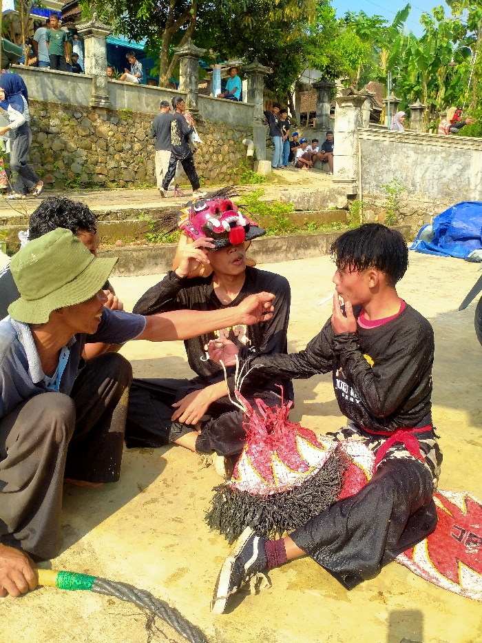 An outdoor scene in Gebyog, Indonesia, captured under bright daylight, features three adult males in the foreground resting on a light-colored ground surface.
The subject on the left, wearing a green bucket hat, a light blue collared shirt, and dark pants, is squatting. His left arm is extended towards the center subject, and he holds a dark, twisted rope or whip with a green-taped wooden handle in his right hand.
The central subject, dressed in a black long-sleeved shirt and dark pants, sits cross-legged. He holds a large, elaborate red, black, and white headpiece or mask, featuring pronounced teeth and a red tongue-like element, positioned over his face, with his gaze directed upward.
The subject on the right, wearing a black long-sleeved shirt with red trim, dark pants, and a red sash, sits with legs extended. He actively smokes a white cigarette held between his right fingers, exhaling smoke. His hair appears wet. Between his legs rests a decorative, woven red and white costume piece with black fringe. His left foot is partially visible, wearing a dark sneaker with white laces and sole.
In the background, an elevated area is defined by a light brown stone wall topped with decorative concrete pillars. Numerous individuals are distributed across this elevated space; some are walking, while others are seated on ledges or steps. Tropical foliage, including banana plants, and parts of light-colored buildings with blue accents are visible further back. A blue tarp is partially visible on the far right, adjacent to what appears to be a motorcycle wheel.