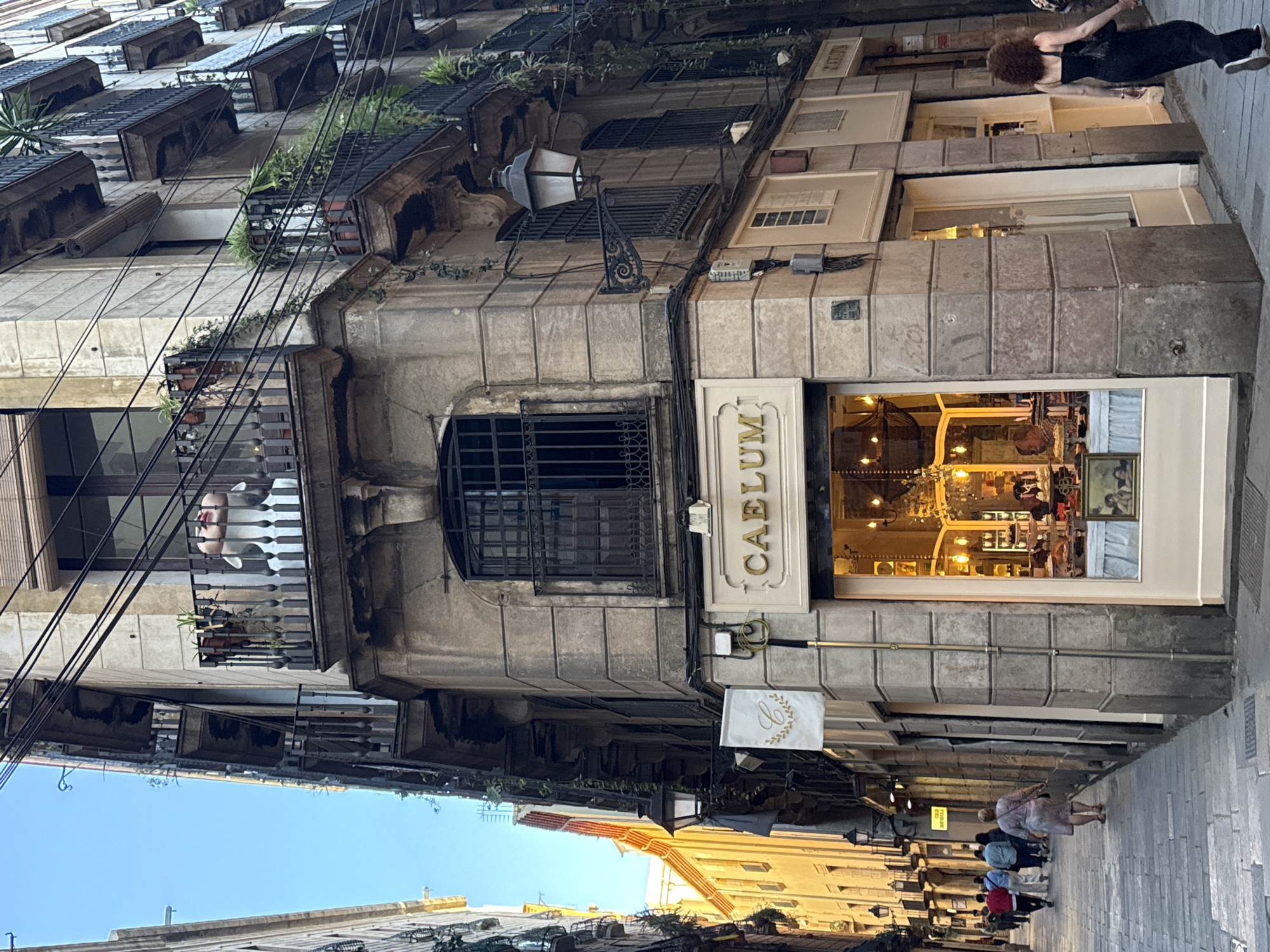 The image depicts a street scene in Barcelona, Spain. The focus is on an old building with a shop called "Caelum" on the ground floor. The architecture is characterized by stone facades, balconies with wrought iron railings, and window boxes with plants. Above the store, a caged window is visible. Power lines run across the facade.
On the right side of the image, a narrow street stretches into the distance, populated with pedestrians. The storefront of "Caelum" is lit from within, displaying goods on shelves. A woman is seen on the sidewalk in the upper right corner of the image, walking away from the camera. Other pedestrians are farther down the street. The sky is visible in the distance between buildings, indicating the time of day might be late afternoon or early evening due to the light.