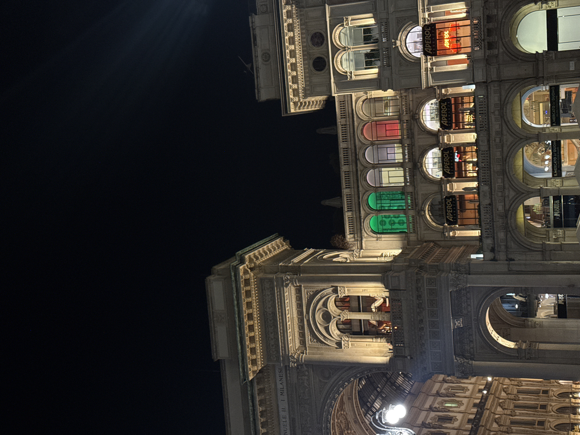 The image shows a night view of the Galleria Vittorio Emanuele II in Milan, Italy. The architecture is illuminated, highlighting its ornate details, including arches, columns, and inscriptions such as "EMANUELE II, I MILANESI." Several establishments display "APEROL" signs in their windows. There is an illuminated facade of a building with various signs with the word "Aperol" on them. The windows above are multicolored. Below is a shopping arcade, with the business “Il Mercato Duomo” visible. Street lamps are visible. The rest of the frame is filled with a dark night sky.