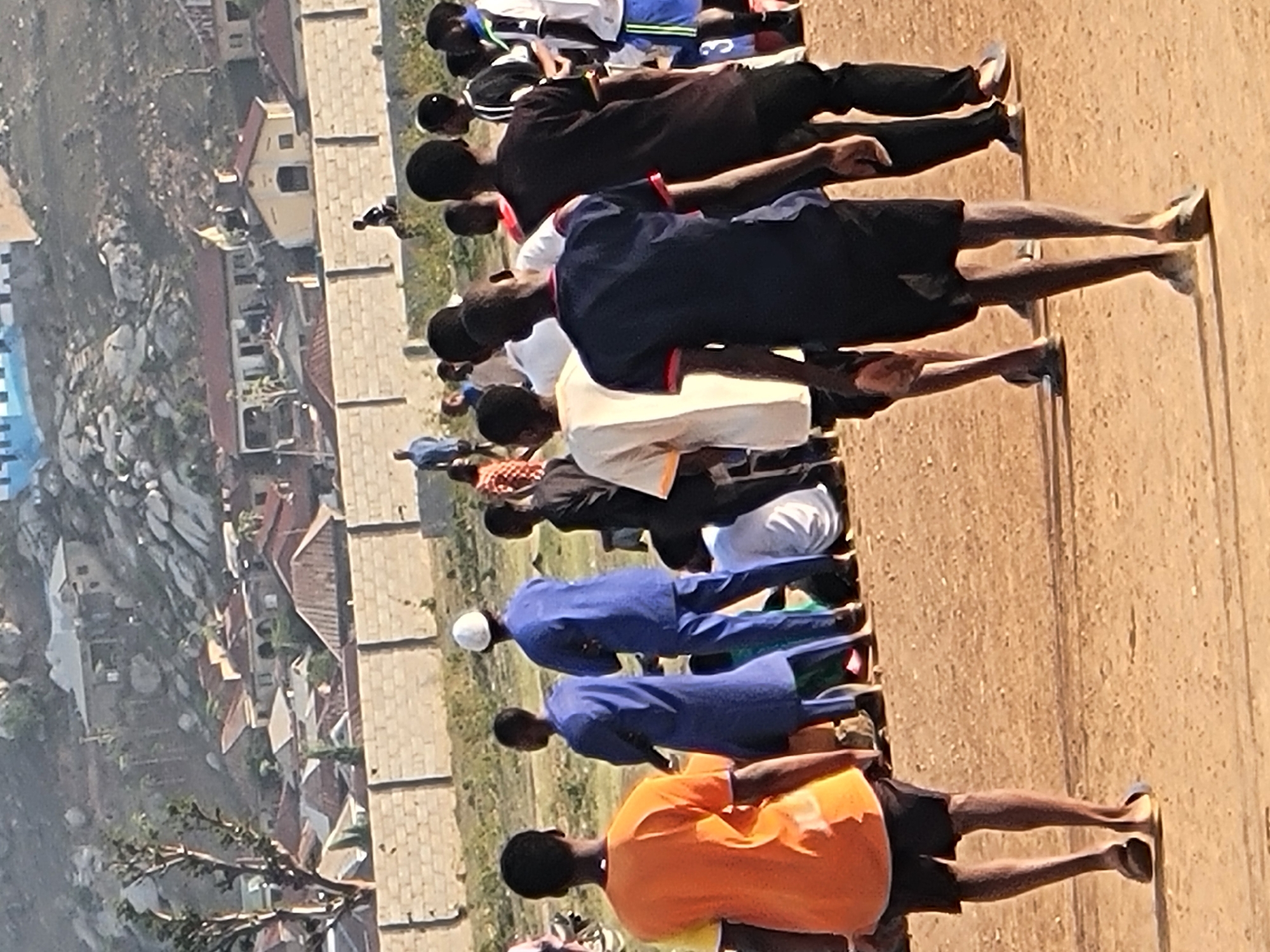 A group of approximately 15-20 people are lined up on a dirt field in Ibo, Nigeria. They appear to be performing some kind of exercise, as they are bending forward from the waist with arms extended. The people are wearing a variety of clothing, including different colors of shirts and pants, and some are wearing head coverings. A brick wall runs along one side of the field, and behind the wall are houses and other buildings. In the background, there is a hillside with rocks and vegetation.
