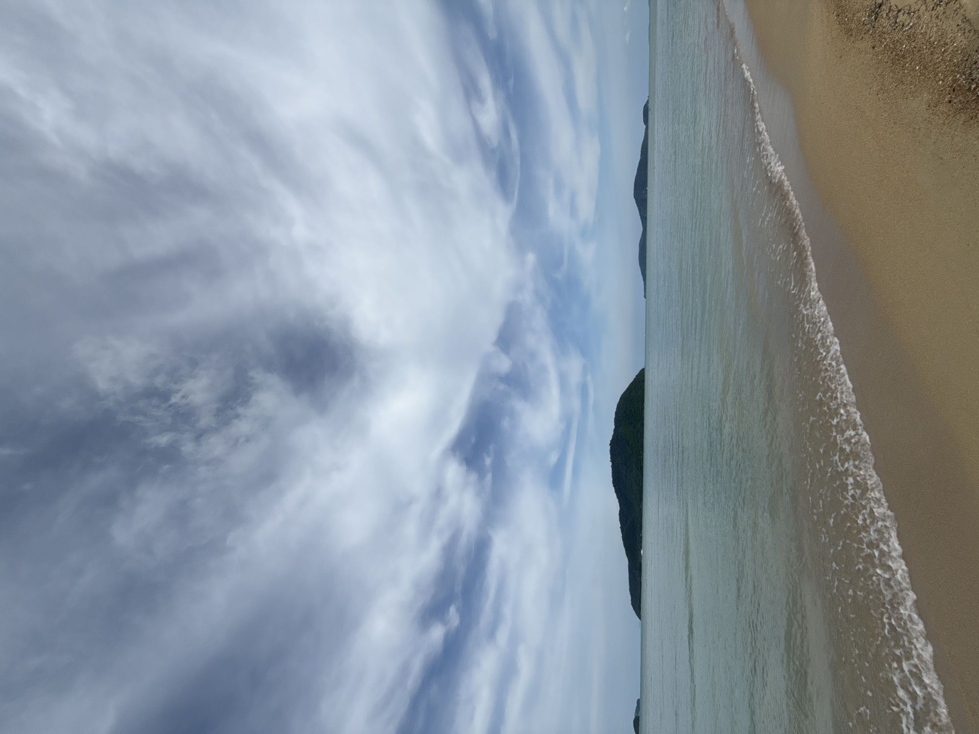 The image shows a serene beach scene at Rawai, Thailand. In the foreground, a gentle wave laps onto a sandy shore, with a small amount of seaweed and shells visible at the edge of the sand. The water is a calm, greenish-blue, stretching out to the horizon. In the distance, several lush green islands rise from the sea, their dark foliage contrasting with the light blue sky. The sky is filled with dramatic, wispy clouds, streaked with shades of gray and white, creating a dynamic and moody atmosphere. The overall impression is one of natural beauty and tranquility. There are no people or animals visible in the image.