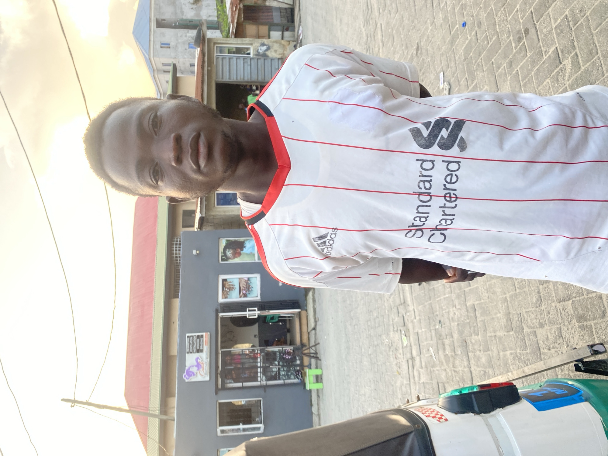 In Ado, Nigeria, a young man stands in front of a building with a red roof. He is wearing a white and red striped shirt with "Adidas" and "Standard Chartered" logos. Behind him, the building appears to be a shop with windows displaying items. A tricycle is partially visible on the left side of the frame. The ground is paved with cobblestones. The sky is cloudy with hints of an overcast sun.