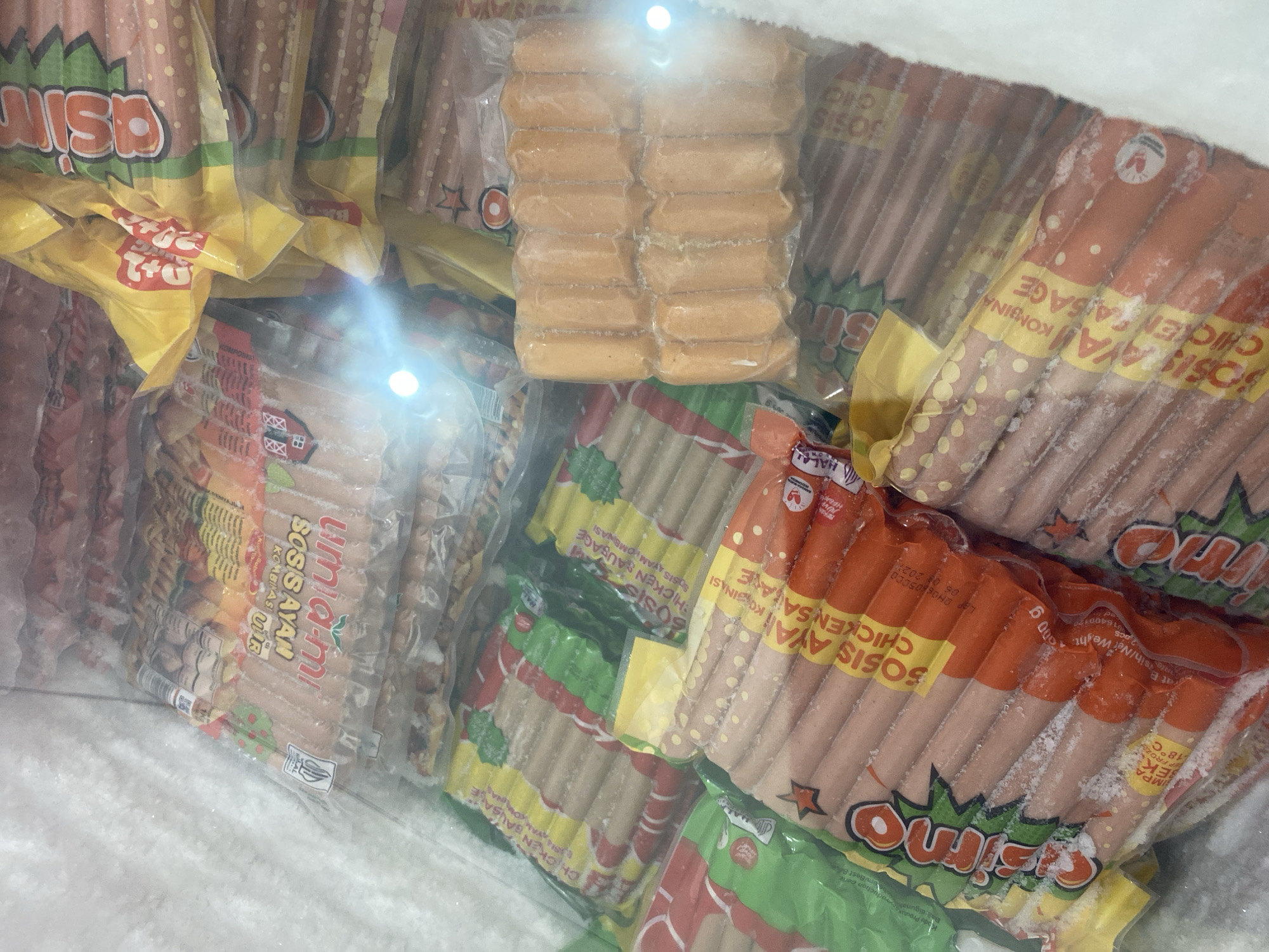 The image shows a freezer filled with packages of chicken sausages. The sausages are packaged in clear plastic with various branded labels in red, green, and yellow. Some visible brand names include "asimo" and "umiami". The packages are stacked and arranged in rows within the freezer. There are no people visible in the image, and no specific location within Payatieng, Indonesia, can be identified. The environment is that of a commercial freezer.