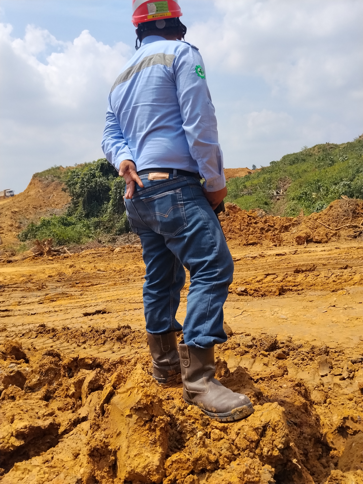 A person wearing a blue long-sleeved shirt, blue jeans, brown work boots, and a red and white hard hat stands on disturbed, orange-brown earth under a cloudy sky. The person's back is mostly to the camera, with their left hand tucked into their back pocket and their right hand obscured behind their back. The shirt has a gray stripe across the back and a green circular logo with a plus sign on the left sleeve. The jeans have a visible brown belt with a rectangular buckle. The boots appear to be made of leather and are dirty. The environment is an outdoor construction or excavation site, with piles of earth, vegetation on hills in the background, and a distant truck on a hill. The ground is uneven and appears to be made of clay or mud.