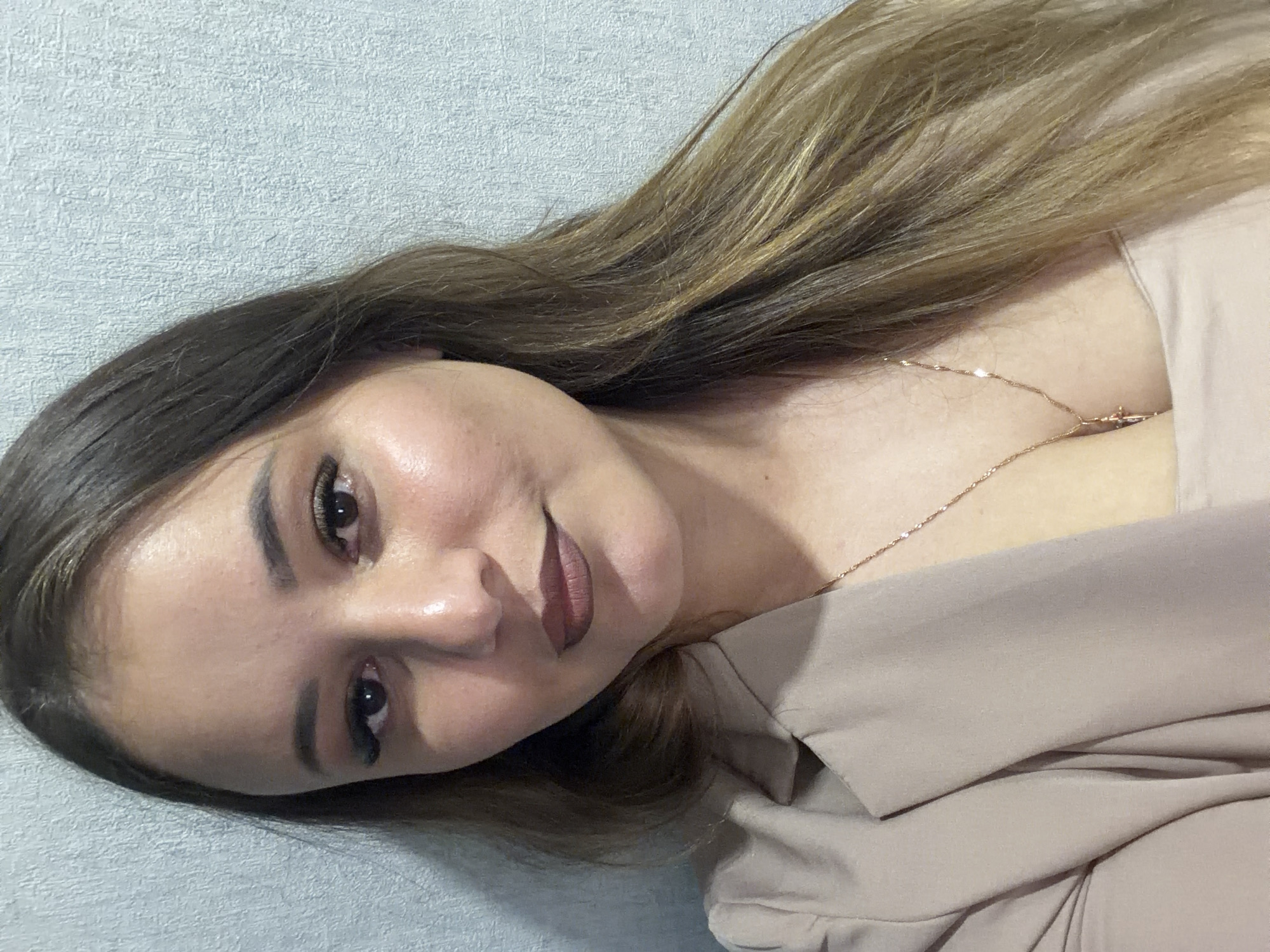 A close-up, chest-up studio portrait shows a woman with long, brown, highlighted hair. She is wearing a tan blazer over a tan top, and a thin gold chain necklace with a cross pendant. Her makeup includes dark eyeliner and mascara, and a brown lipstick. The background is a textured, light blue wall. There is no indication of the specific location within the city of Chaikovsky, Russia.