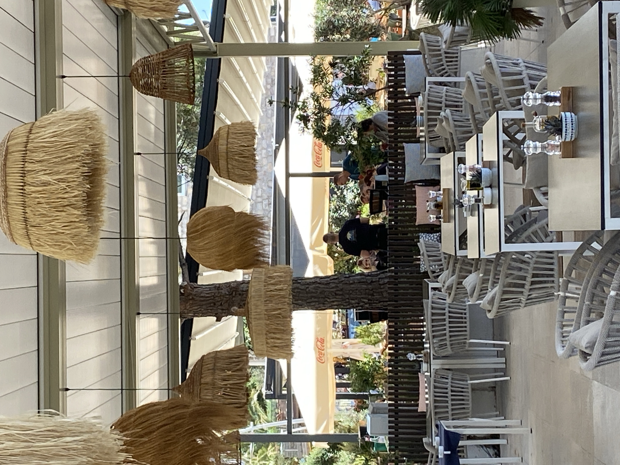 The image depicts an outdoor seating area of a cafe or restaurant in Petrovac centar, Montenegro. The scene is set under a white retractable roof, with several large, straw-like lamps hanging from above. The foreground is dominated by tables and chairs. Most of the chairs are woven with a light-colored material, featuring grey cushions. The tables have metallic surfaces and white legs. On some tables, salt and pepper grinders, small potted plants, and oil dispensers are visible.
In the background, people are visible at various tables. One table features a Coca-Cola umbrella. A man in a dark t-shirt is standing, and another man in a blue t-shirt is leaning forward, seemingly interacting with someone at a table. Several other individuals are seated at tables, some partially obscured by foliage and other structures. The environment appears to be lush with greenery, with trees and bushes surrounding the outdoor seating. The overall impression is that of a relaxed, open-air dining establishment.