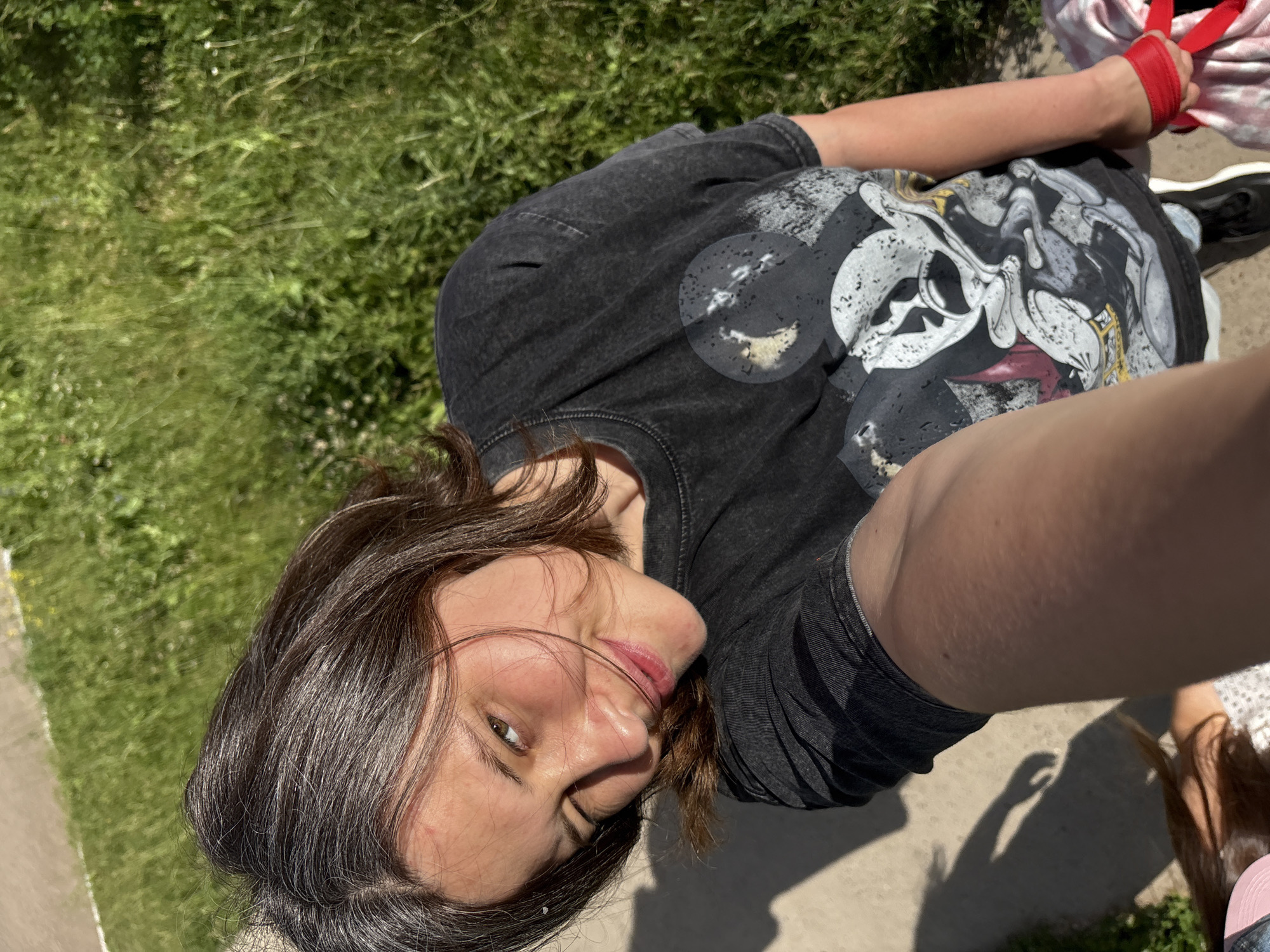 A young woman with brown hair, wearing a dark t-shirt with a skull print, takes a selfie outdoors. She is lying down, with her face close to the camera and her eyes looking into the lens. Her expression is relaxed and happy, with a slight smile. The background shows green grass, suggesting a park or a grassy area. The lighting indicates it is daytime, likely sunny. The woman's arm is visible, and she is wearing a red band on her wrist. Another person's hand and arm are also visible in the upper right corner, wearing a similar red band. The scene appears casual and informal, possibly a selfie taken with a friend or companion. The location is specified as Chaikovsky, Russia.