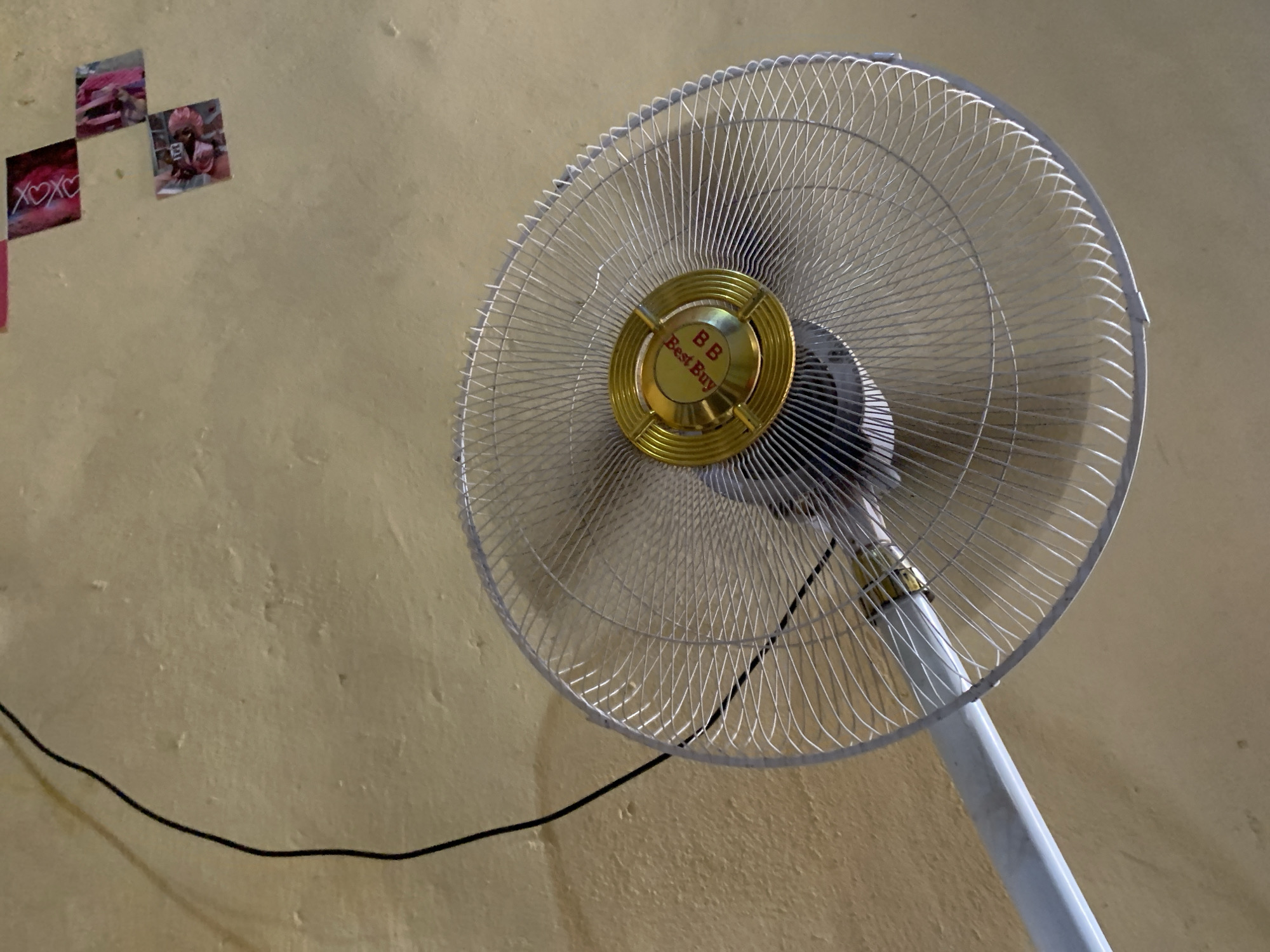 A standing fan dominates the scene, positioned in front of a pale yellow wall. The fan has a silver-colored wire cage and a golden center with "BB Best Buy" printed on it. A black power cord extends from the fan and runs along the lower left portion of the wall. On the upper left of the wall, there are a few small square images or photographs. The environment appears to be indoors. The image was taken in Keffi, Nigeria.