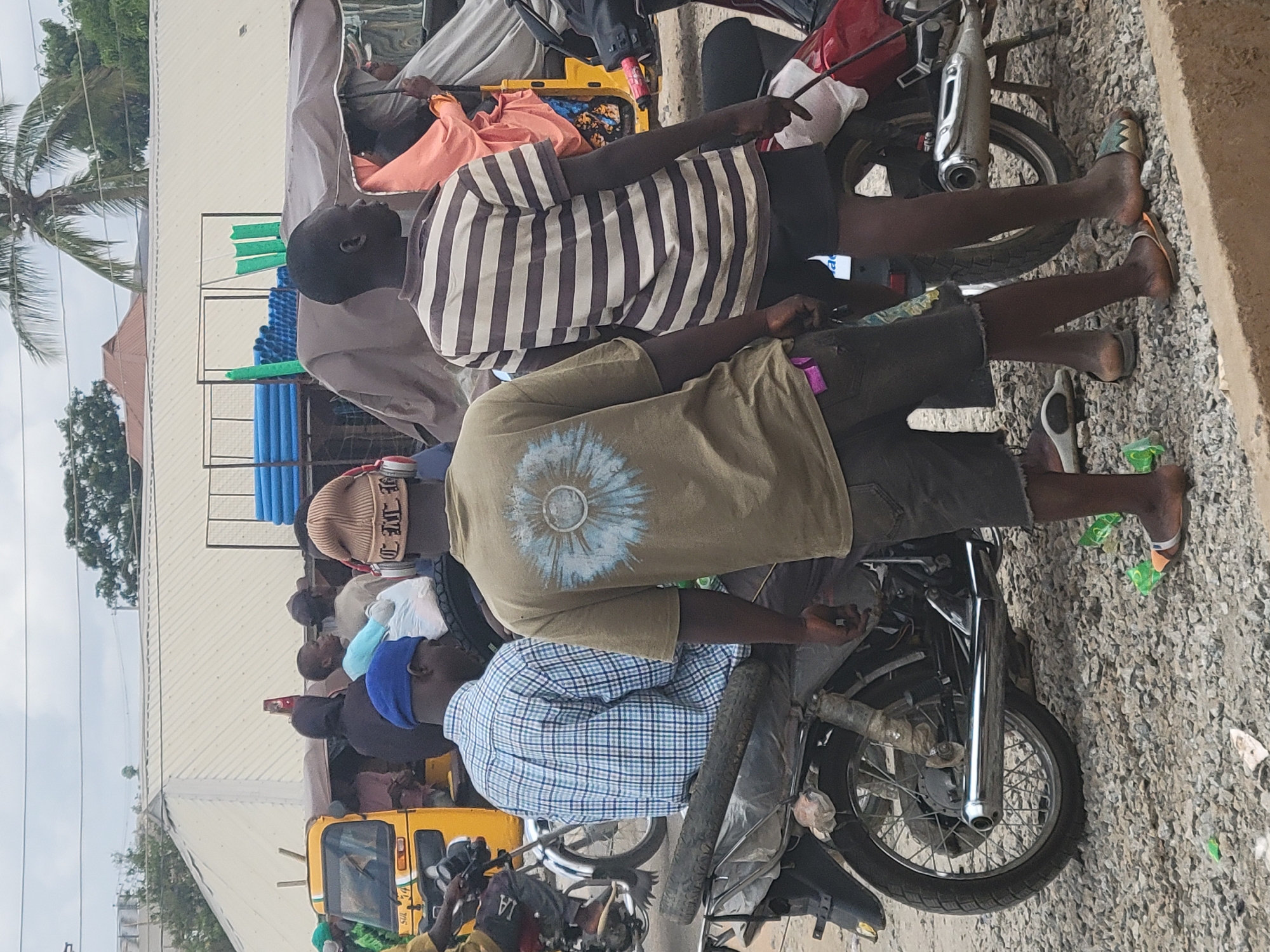 The image shows several people sitting on motorcycles in Suleja, Nigeria. The people are wearing casual clothing. One man wears a striped shirt, another a green shirt with a sun pattern on the back, and a third wears a blue plaid shirt. The motorcycles are parked on a street with gravel and trash. Other people and vehicles, including a yellow auto rickshaw, are visible in the background. Blue and green pipes are also being transported on one of the motorcycles.
