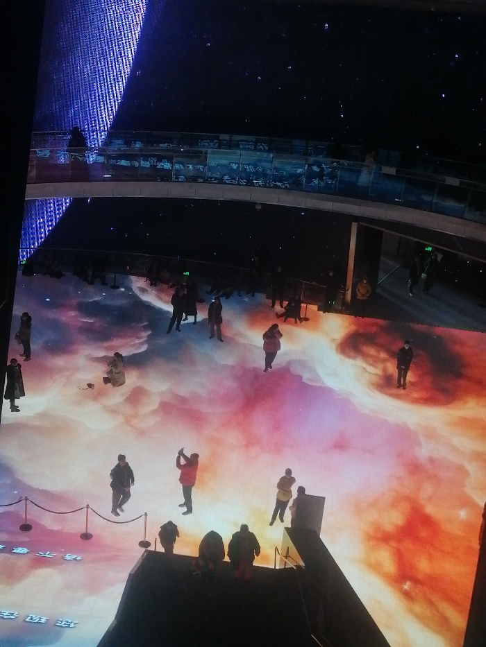 The image captures an expansive, dimly lit interior public exhibition space within Fengtai District, China, dominated by a large-scale, interactive floor projection. The projection vividly displays cosmic imagery, featuring swirling nebulae in gradients of pink, orange, purple, and white, which transition into a dark, star-filled expanse. Approximately 20 to 25 individuals are present on the projected floor, primarily standing and observing. One person in a red jacket, located near the bottom-center, is actively holding a device, likely a smartphone, upwards as if photographing or videoing the scene. Another individual, dressed in light-colored attire, is crouched on the mid-left section of the projection. A dark barrier, composed of stanchions and ropes, delineates a section of the floor on the lower-left.
Above the main projected floor, a curved, multi-level walkway with glass railings spans the upper portion of the frame; one person is visible on this walkway to the far left. The upper-left background features a tall, vertical blue illuminated surface with a textured appearance, while the rest of the upper area is dark, punctuated by small, scattered lights resembling distant stars. A dark, geometric structure, appearing to be a platform or set of stairs, occupies the lower right foreground, with several individuals positioned on or near it. Further individuals are visible as dark silhouettes on a lower viewing platform or seating area located between the main floor and the upper walkway.